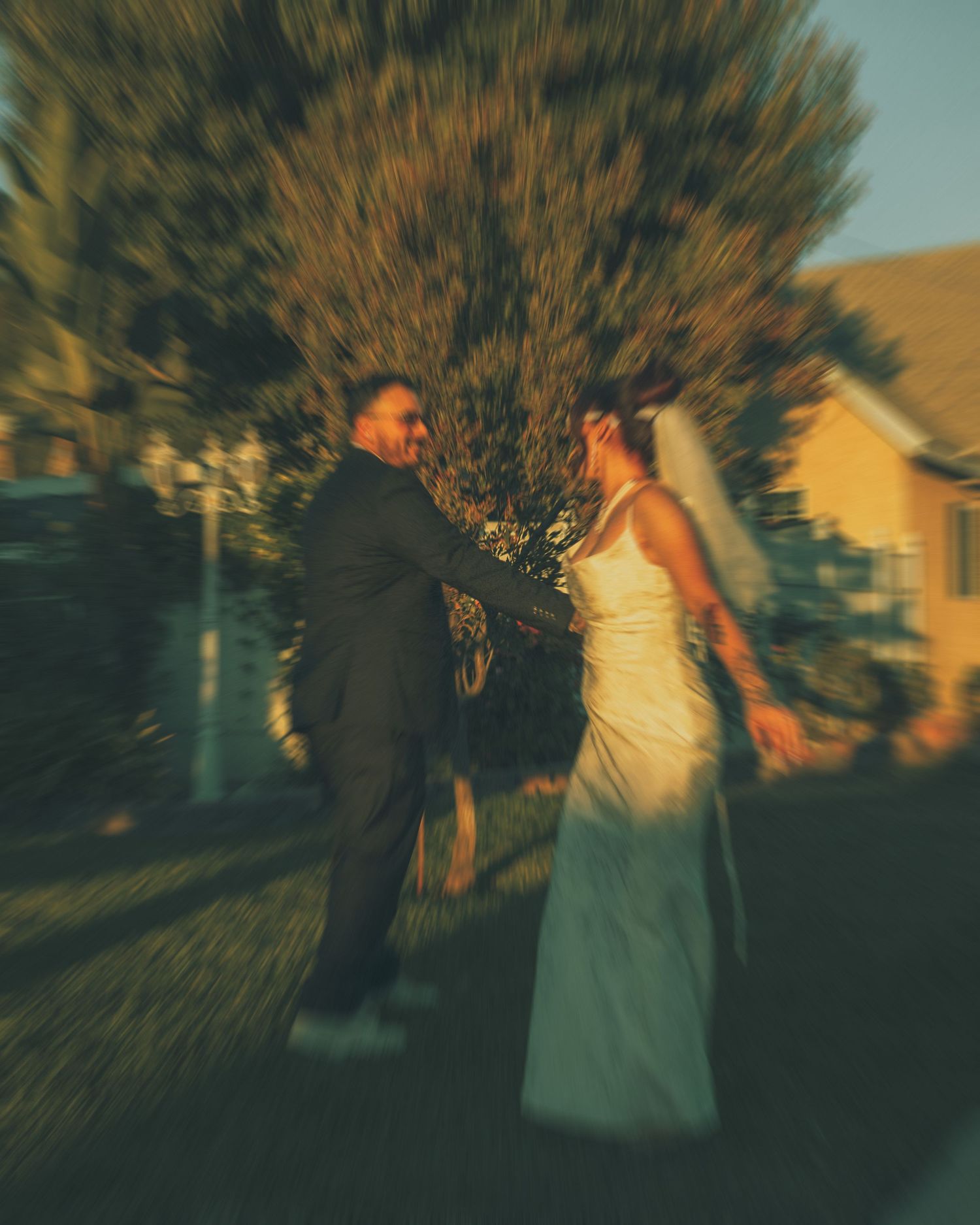A couple in formal wedding attire share a romantic moment outdoors at sunset with a dreamy blurred effect.