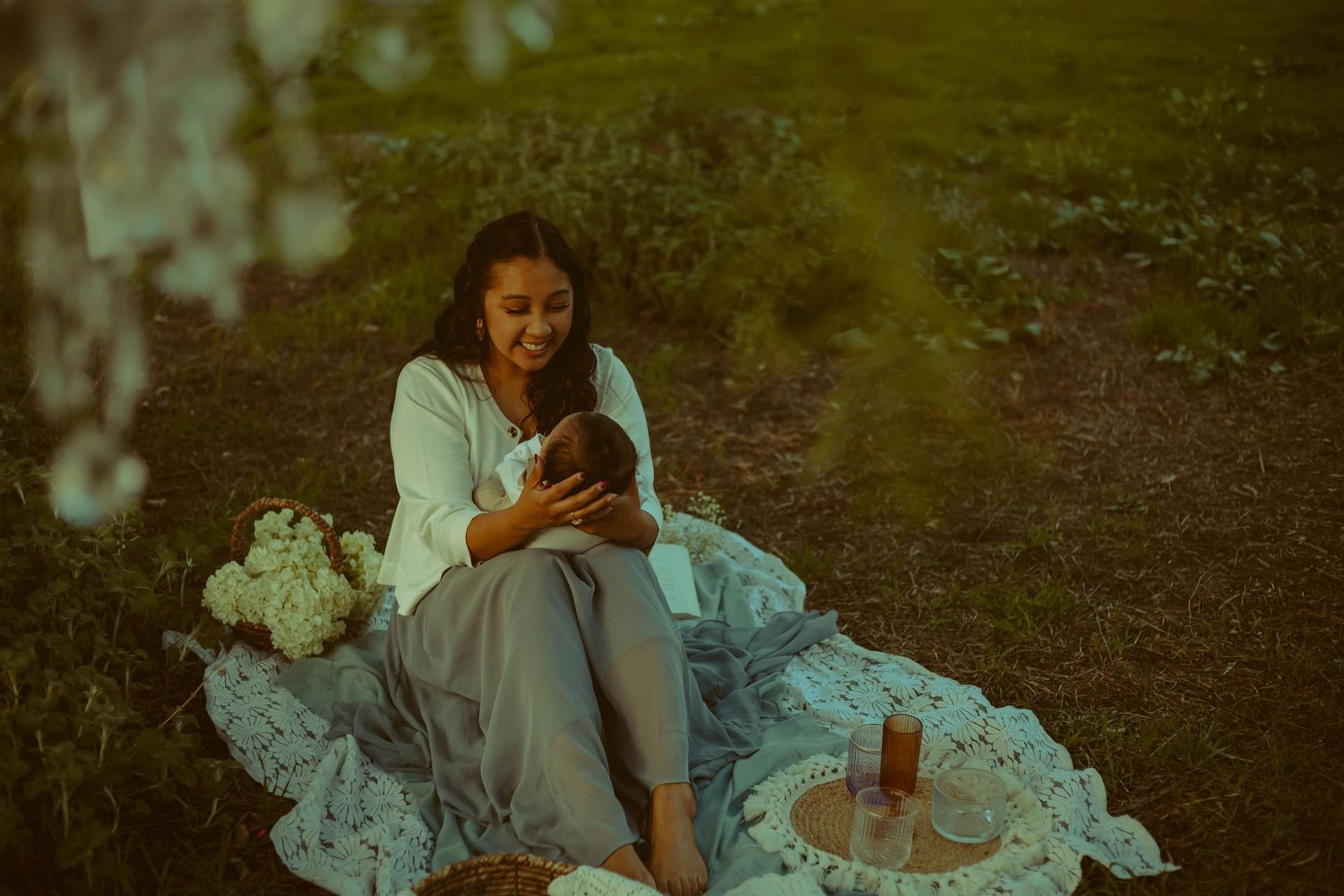 A California mother relaxes on a white lace blanket in a garden setting while playing with her baby during a golden hour mini session.