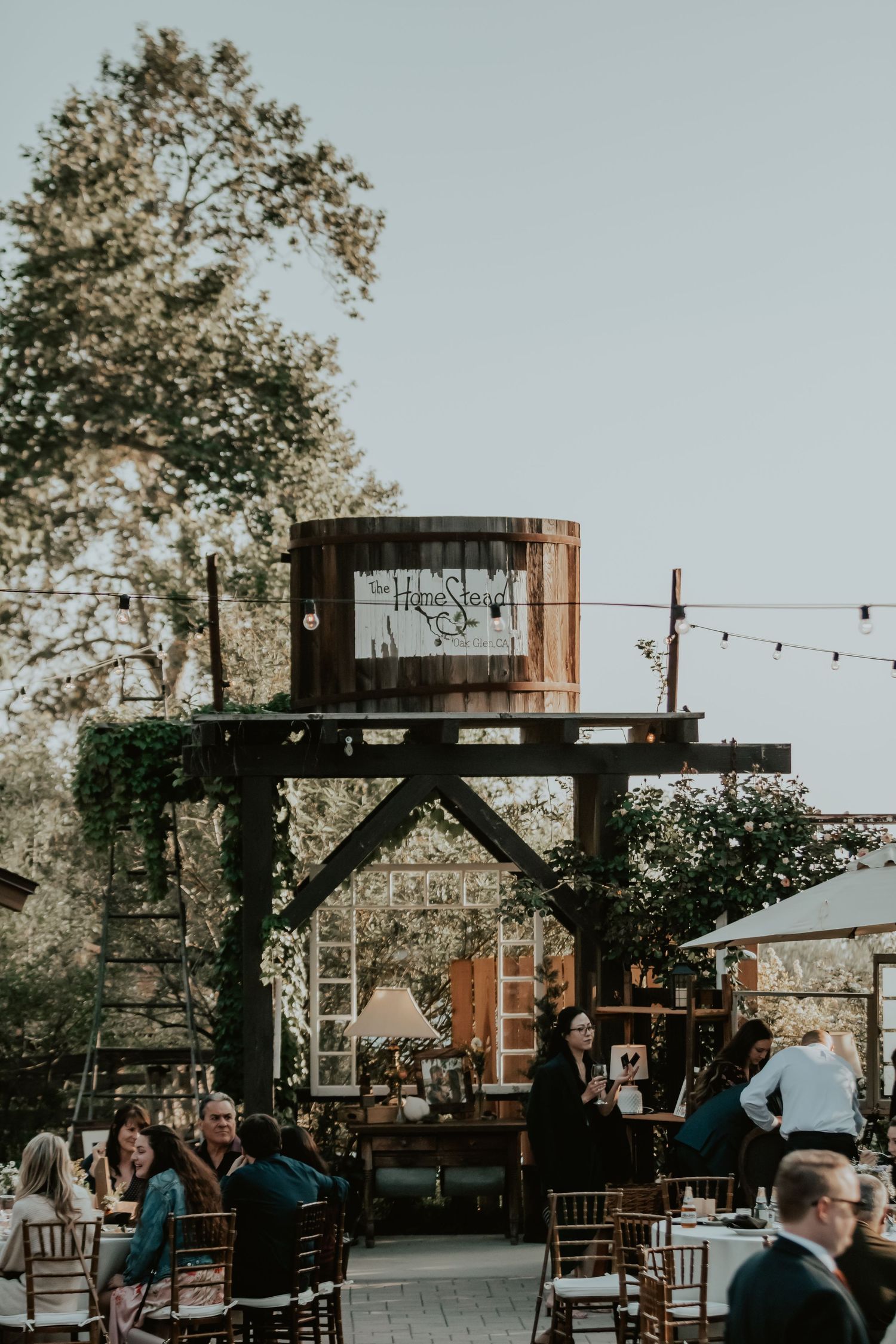 Rustic outdoor wedding venue with wooden water tower, string lights and chairs setup for gathering at dusk.
