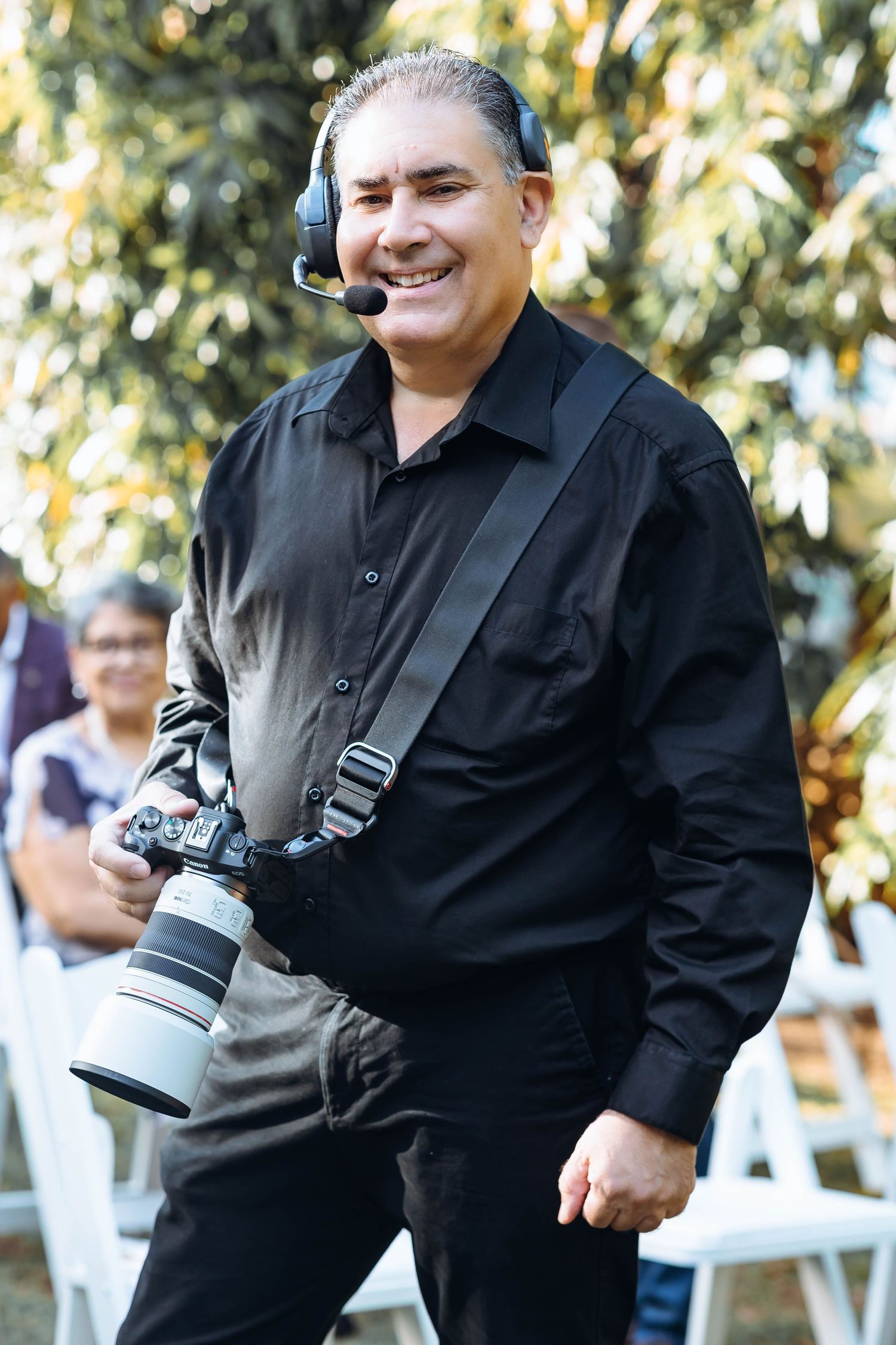 Professional photographer in black attire smiling while carrying camera equipment outdoors.