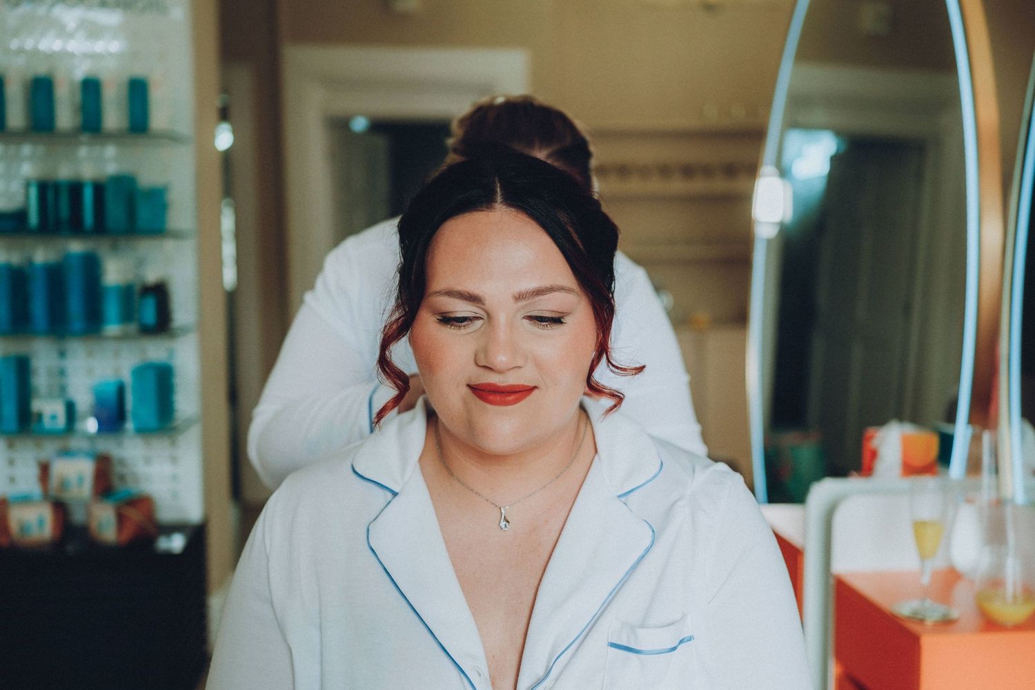 Someone wearing a white satin robe smiles while getting ready in a salon with beauty products in the background.