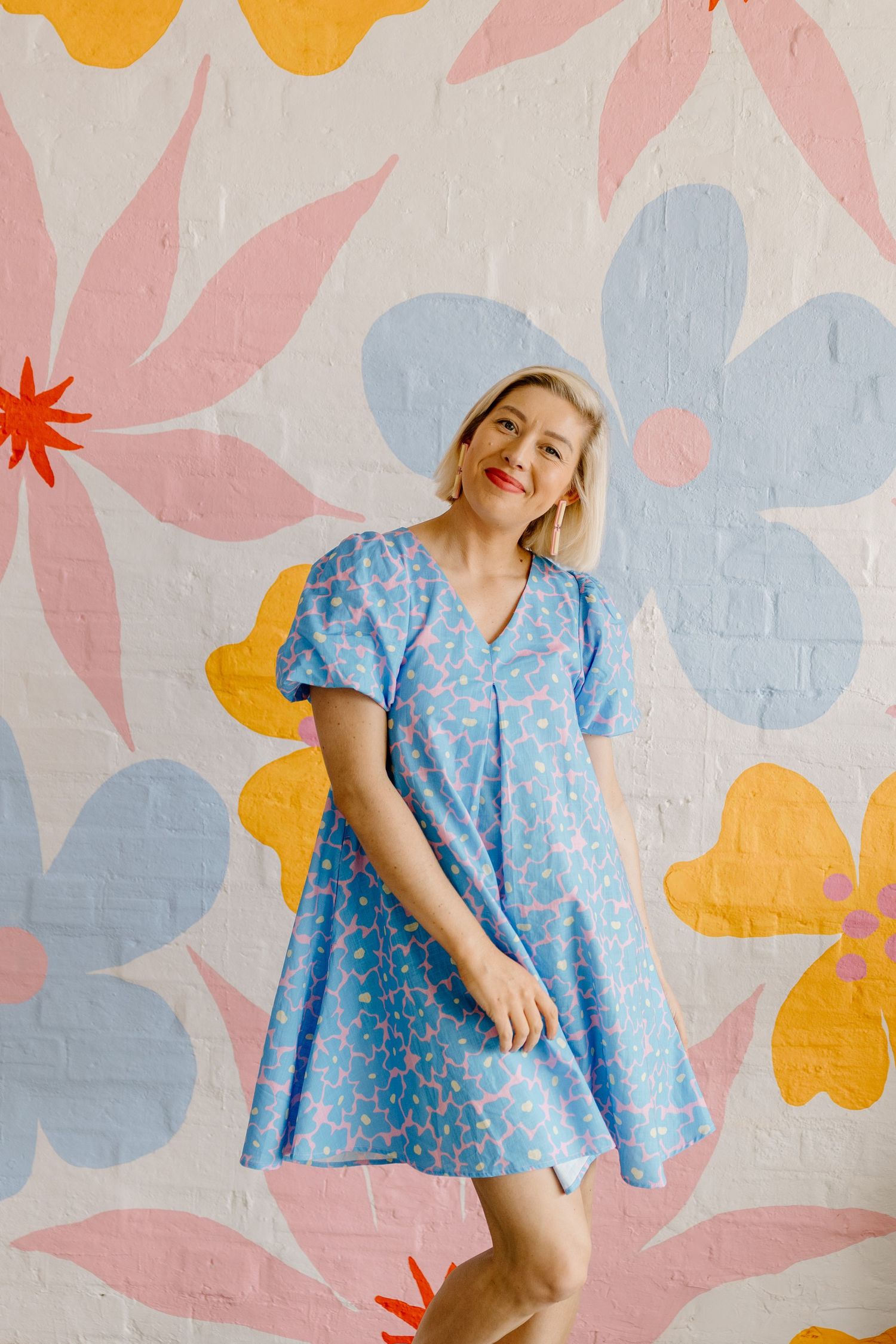 Someone wearing a light blue floral dress poses against a colorful floral wall mural with pink, yellow and orange blooms.