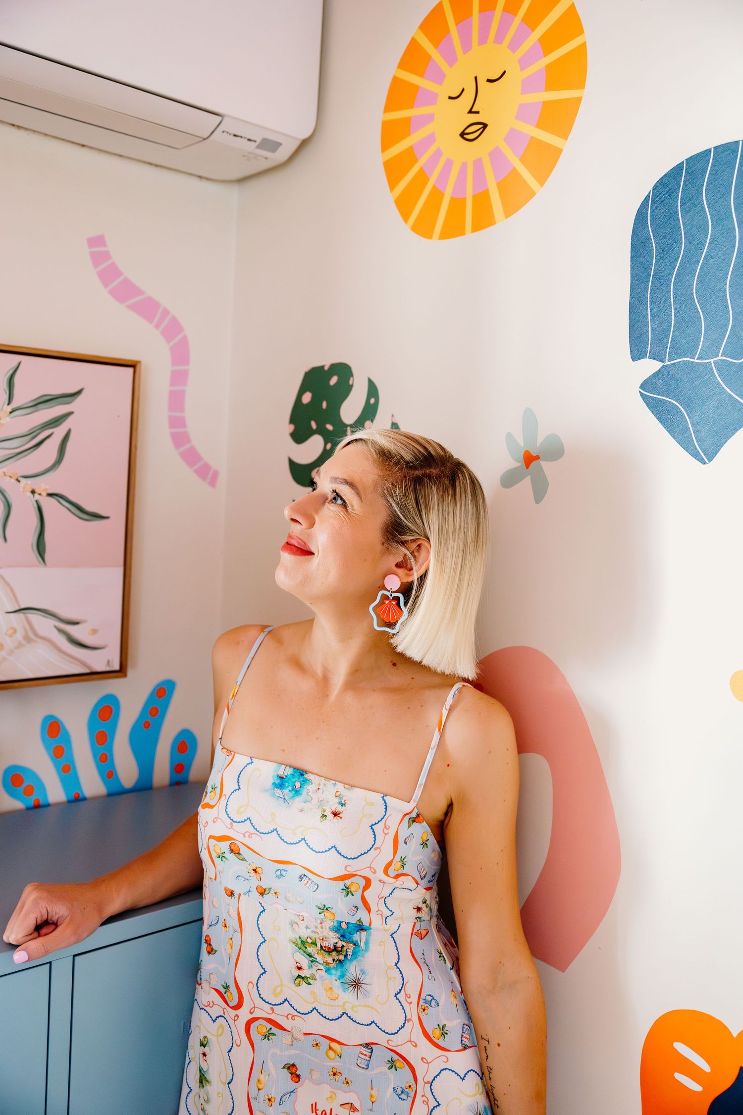 Colorful wall art featuring abstract shapes and sun design decorates room with blonde woman in floral dress laughing.