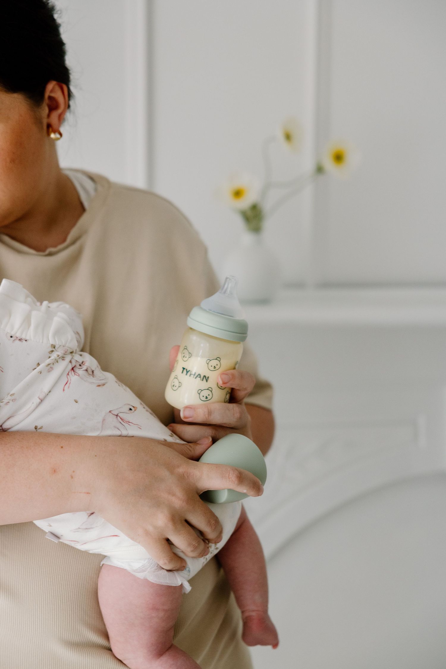 A parent holds a baby in white clothes while feeding from a bottle, with yellow flowers in a vase in the background.