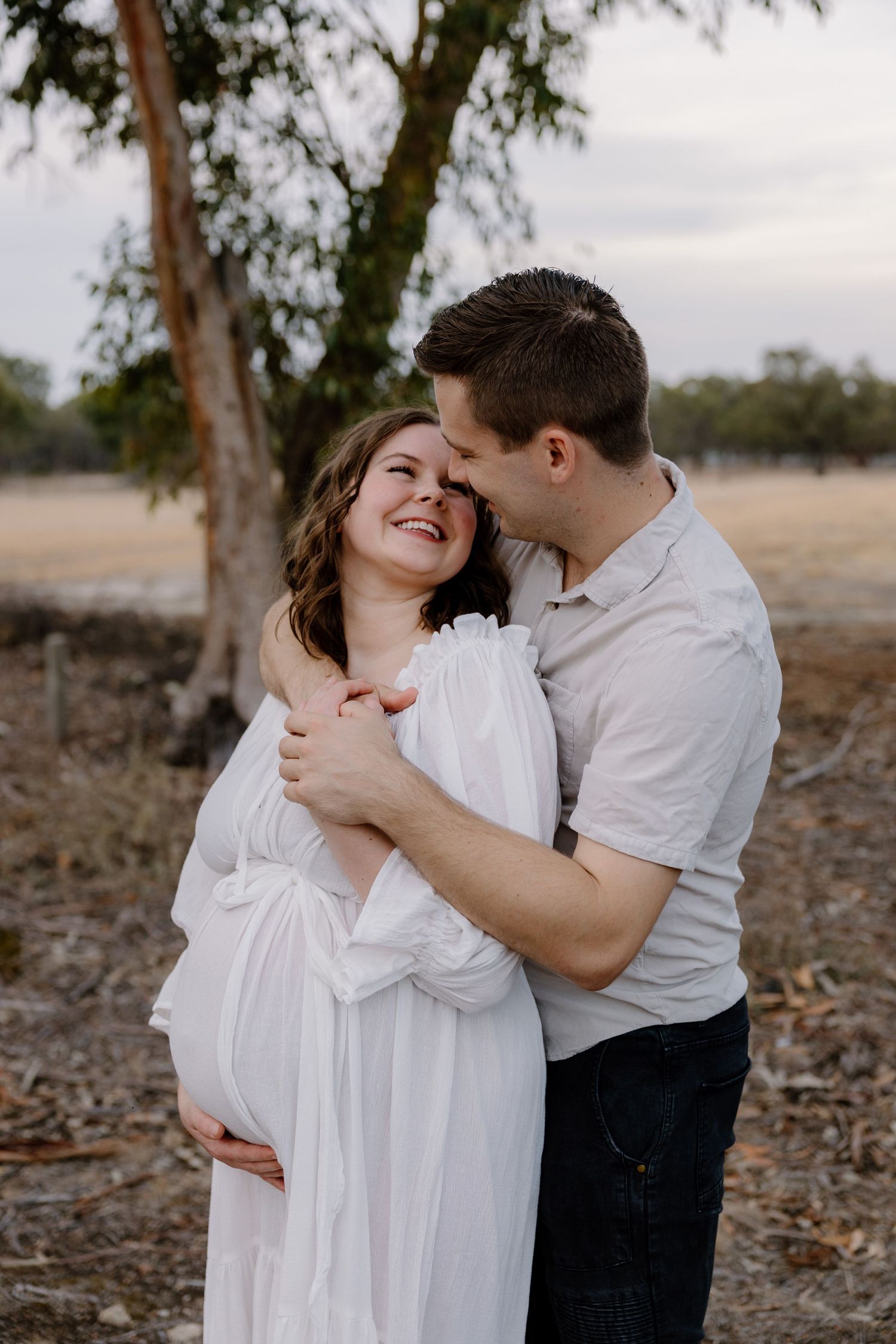 A couple in white clothing shares tender moments together while embracing outdoors among trees during a photo session.