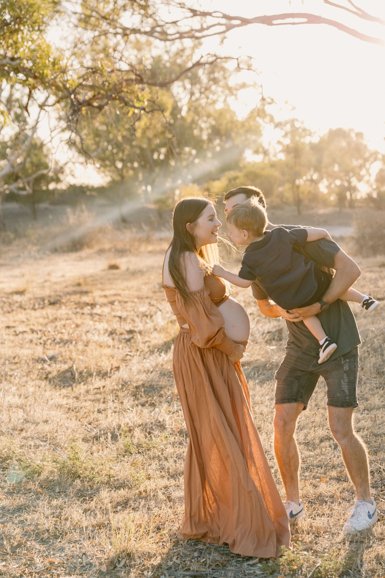 Family stands together in warm sunlit field during golden hour with flowing maxi dress and casual attire.