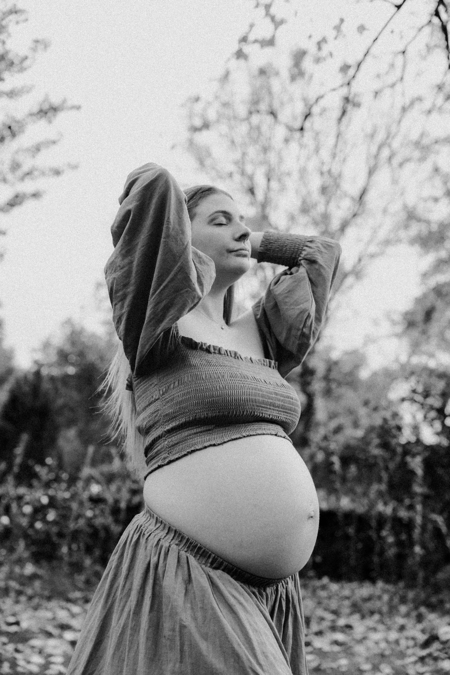 Black and white portrait of expectant mother in a flowing dress stretching outdoors among autumn trees.