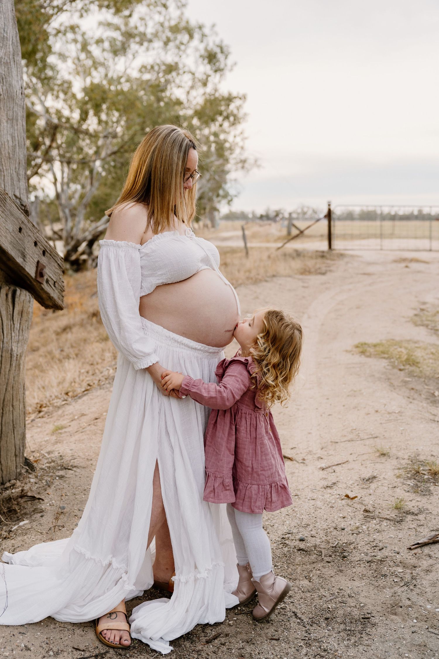 A small child in a pink dress embraces a pregnant woman wearing a flowing white dress on a dirt path at sunset.