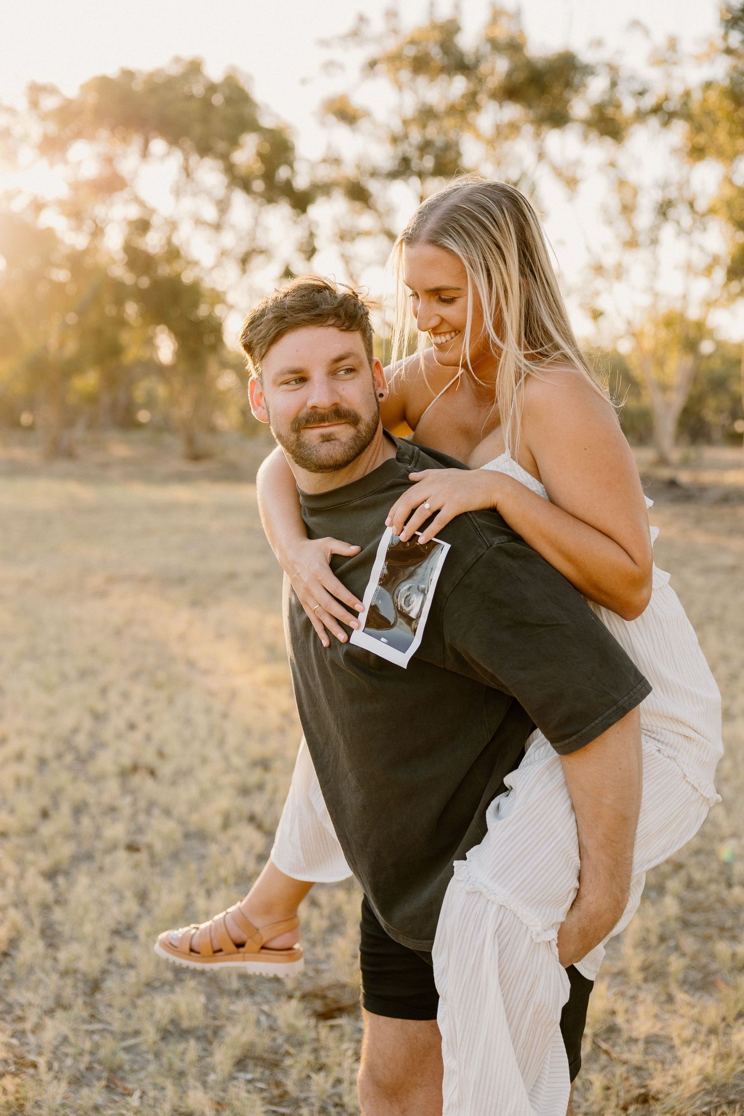 A couple shares a joyful moment in a sunny field during golden hour as he gives her a piggyback ride.