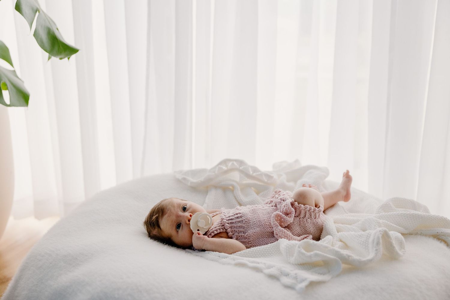 A baby in pink clothing lies contentedly on a white bed near a bright window with flowing curtains.