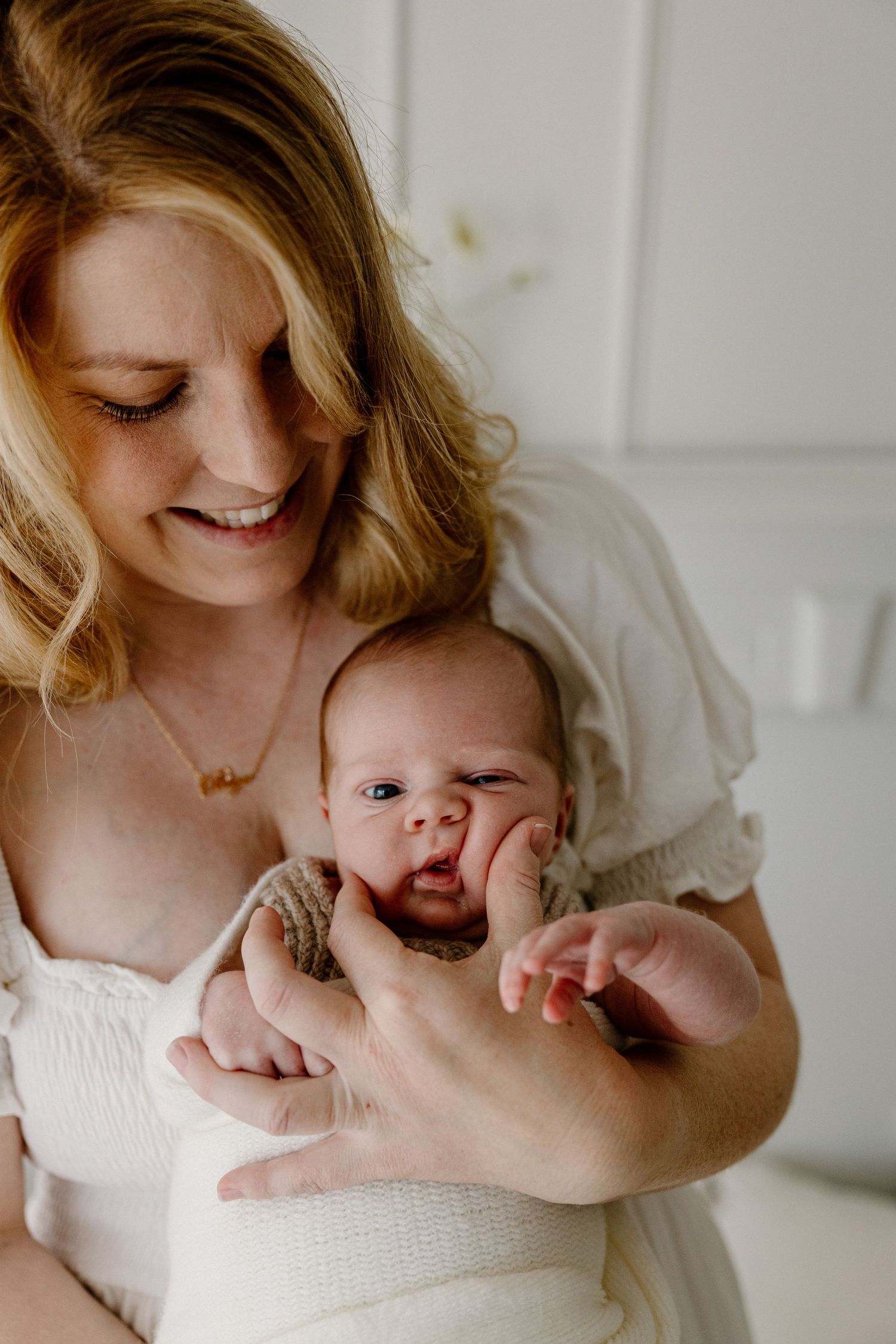 A tender moment captured as parent in white dress cradles a small infant with expressive face in a bright airy setting.