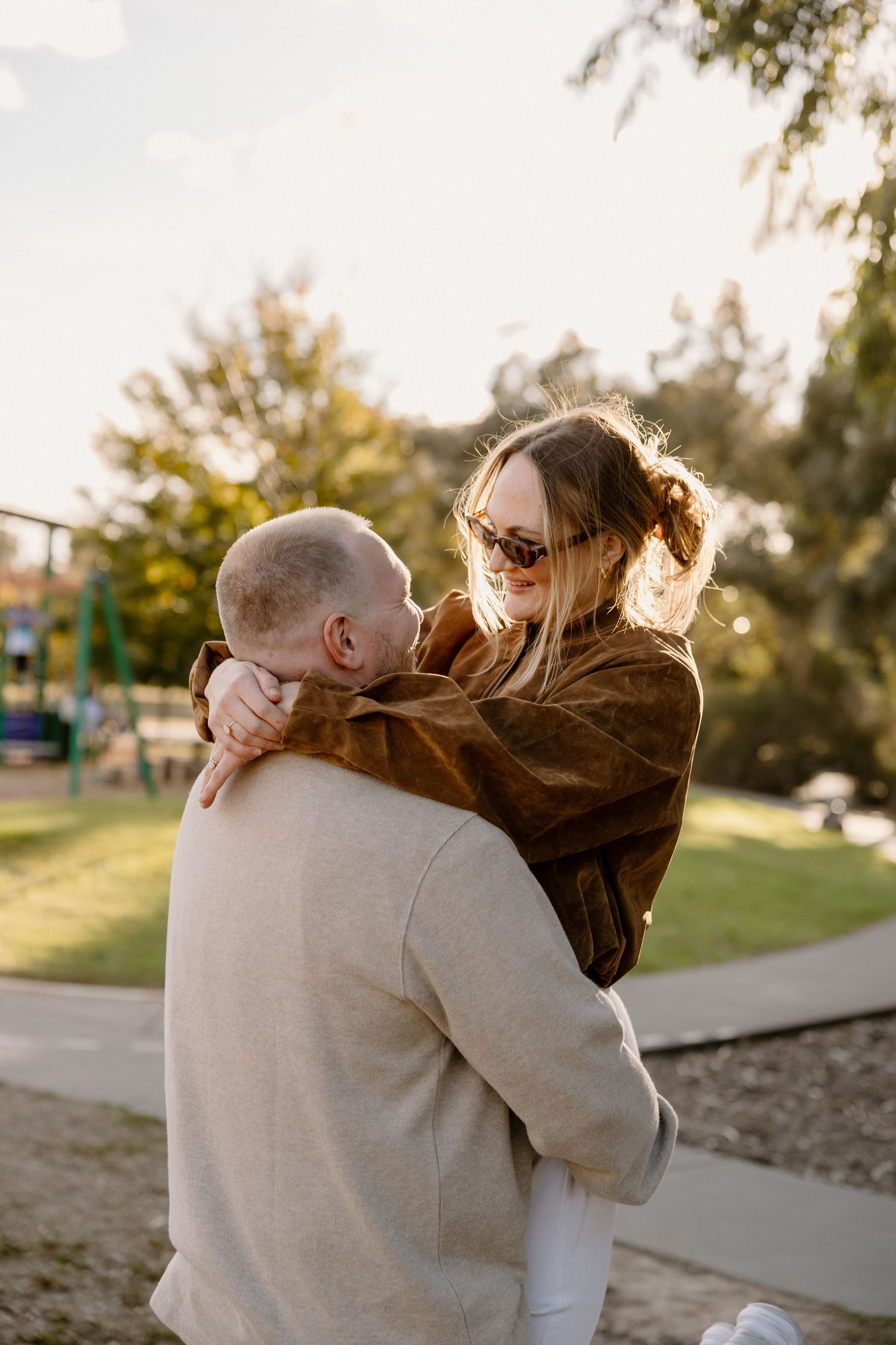 A couple embraces outdoors in a park at sunset, with golden light filtering through trees in the background.