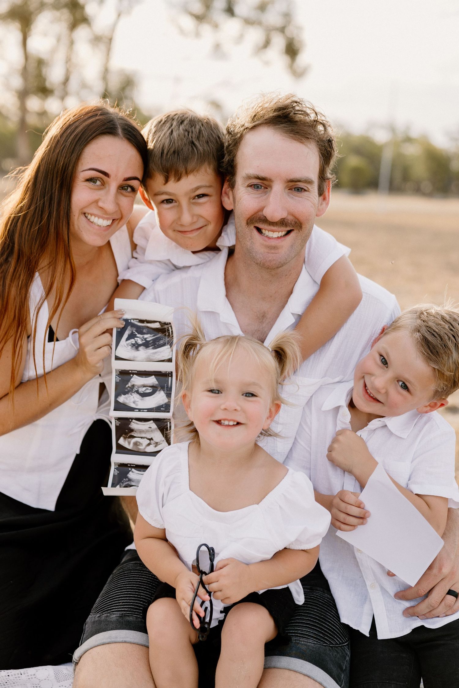 A happy family dressed in white and black outfits share joyful moments while holding an ultrasound photo outdoors.