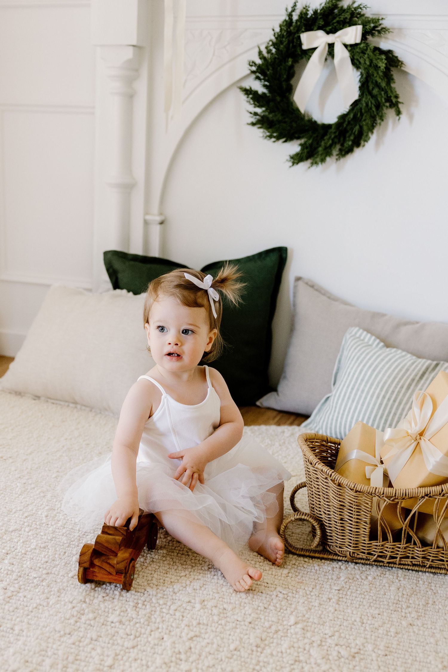 A young child in a white tulle dress sits on carpet beside a basket of wrapped presents and holiday wreath on wall.