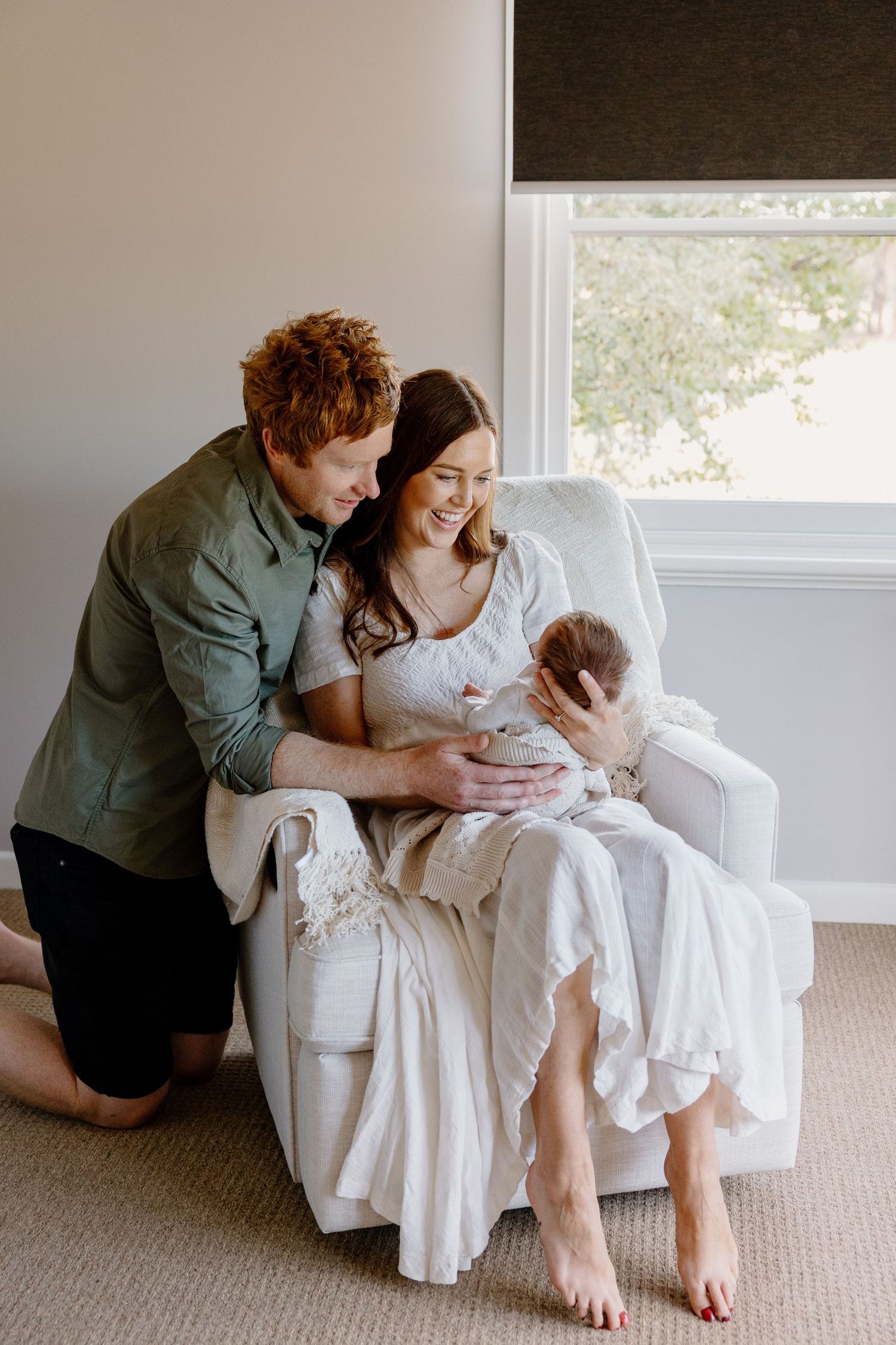 A couple sitting in a white armchair near a window shares an intimate moment together as they embrace their infant.