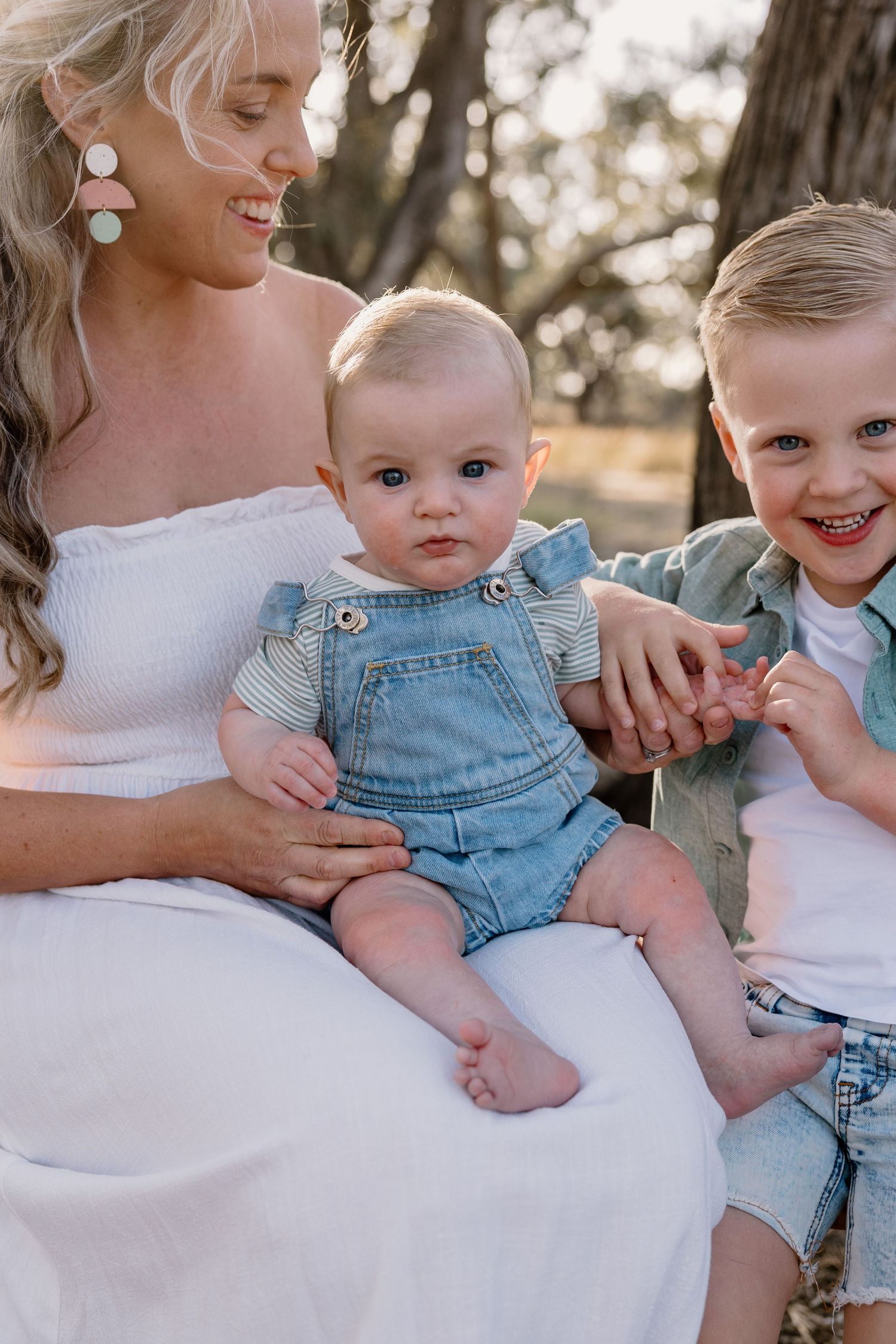 Person in white dress sits outdoors with two children, including baby in denim overalls and toddler in casual wear.