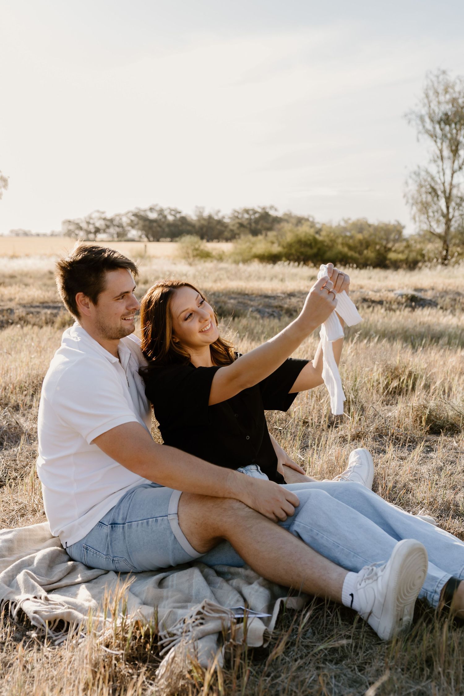 A couple in matching white and denim outfits lounges playfully on golden grass during a sunset photoshoot.