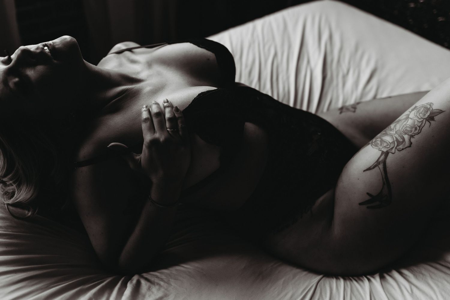 Dramatic black and white photograph of someone resting on white bedding with visible tattoos and shadows.