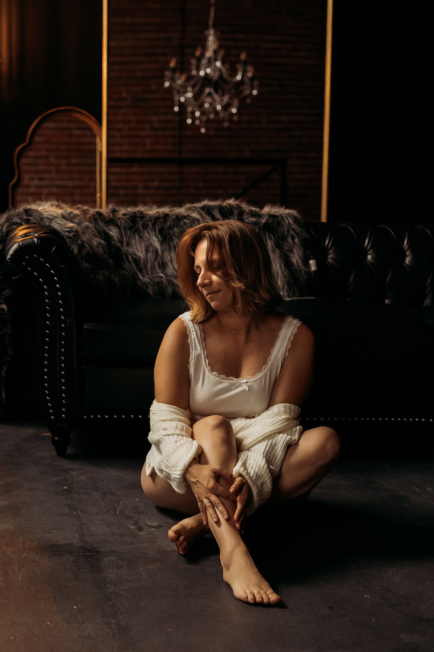 A person in a white tank top and cardigan sits casually on a dark leather couch with an ornate chandelier in the background.