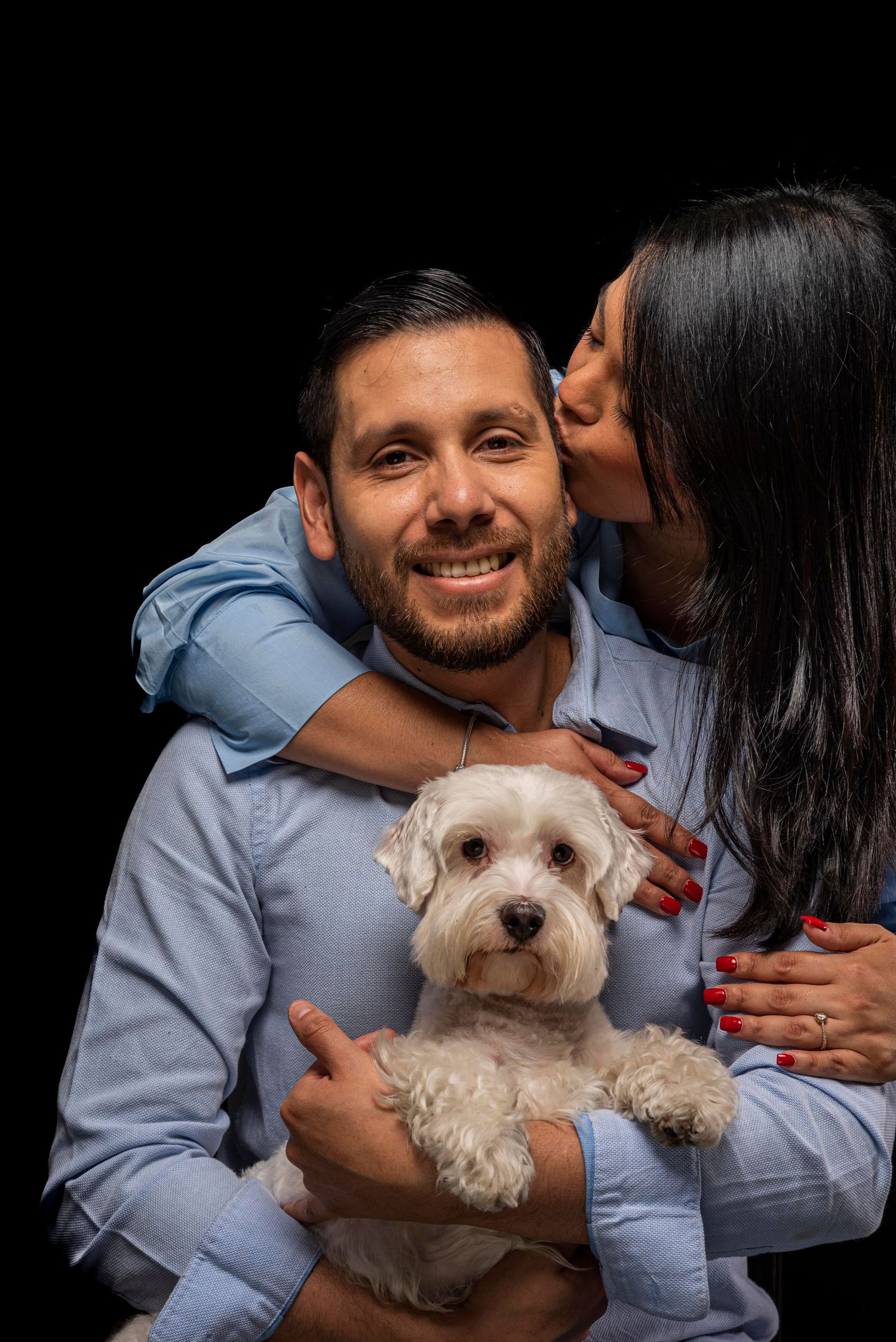 A couple embraces while holding their small white fluffy dog against a black background.
