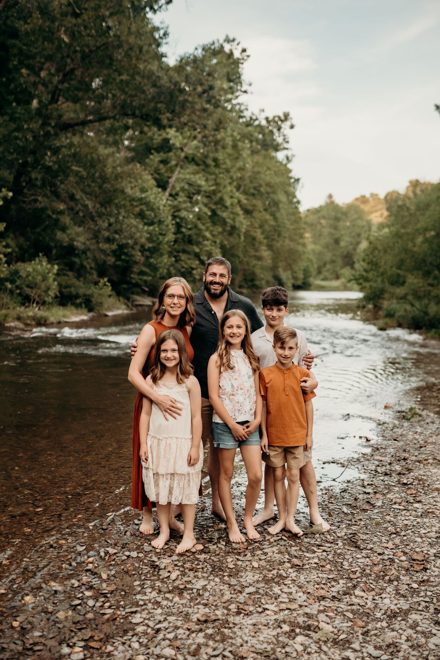 A group in casual attire stands together on a rocky riverbank surrounded by trees on an autumn day.