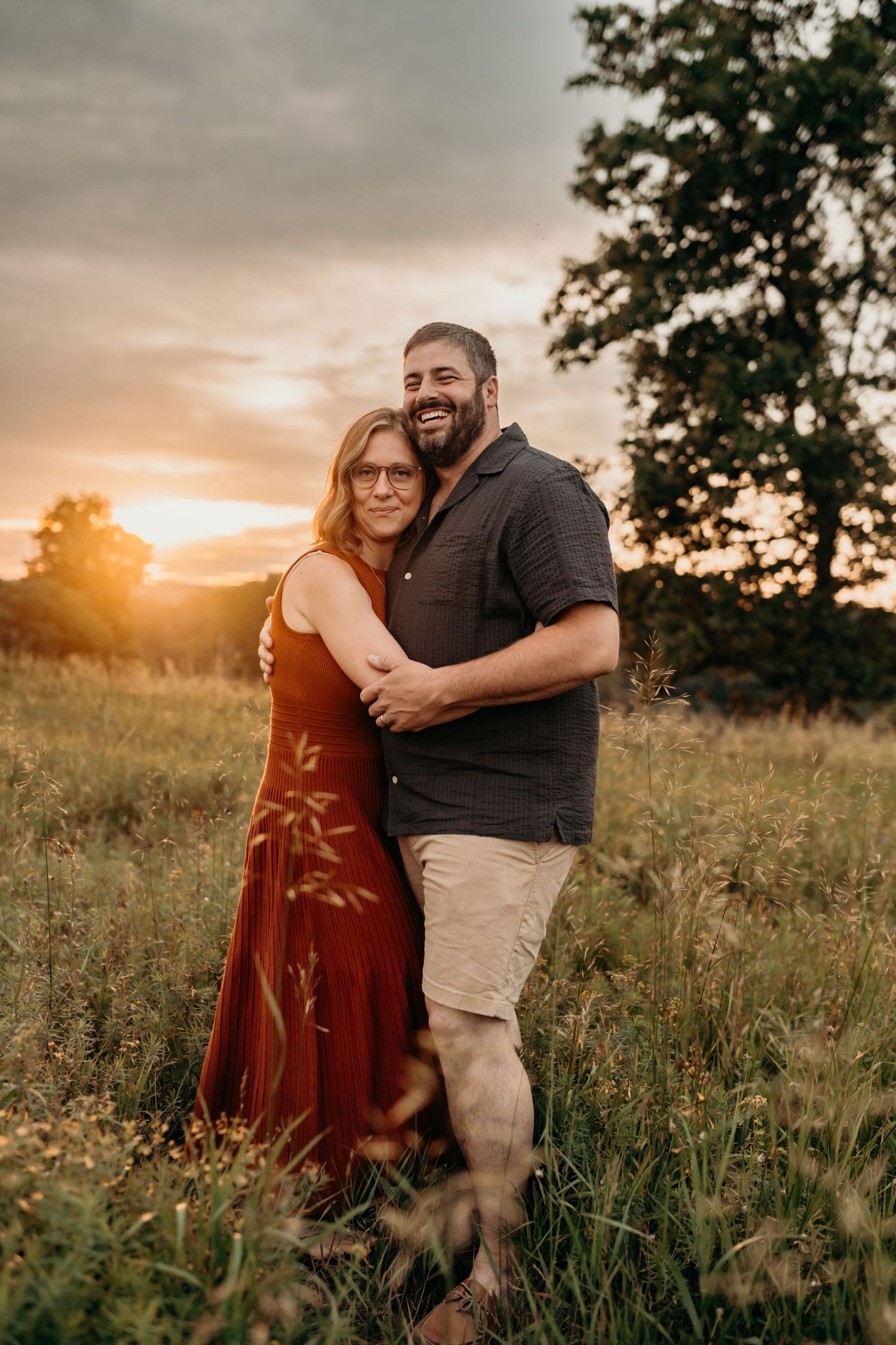 A loving couple embraces in a golden field during sunset, with her wearing a rust-colored dress and him in dark casual attire.