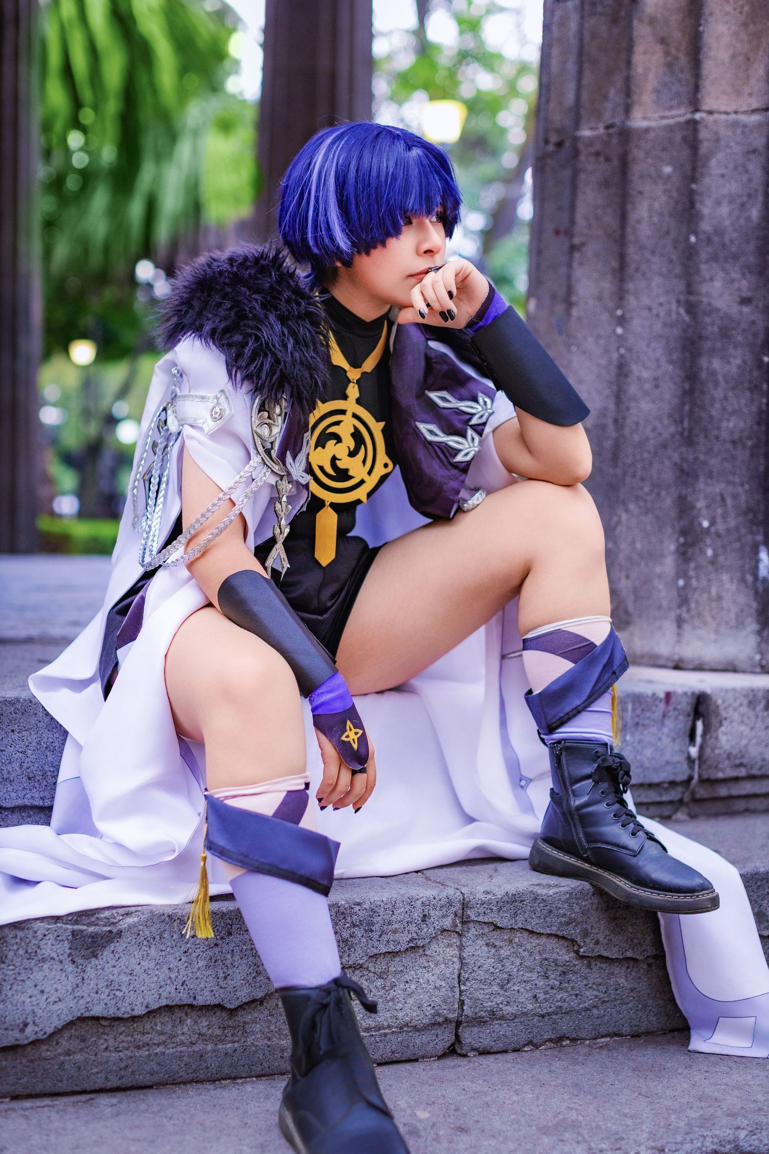 Cosplay photoshoot featuring character with blue hair wearing a white cloak and black top with gold pendant on stone steps.