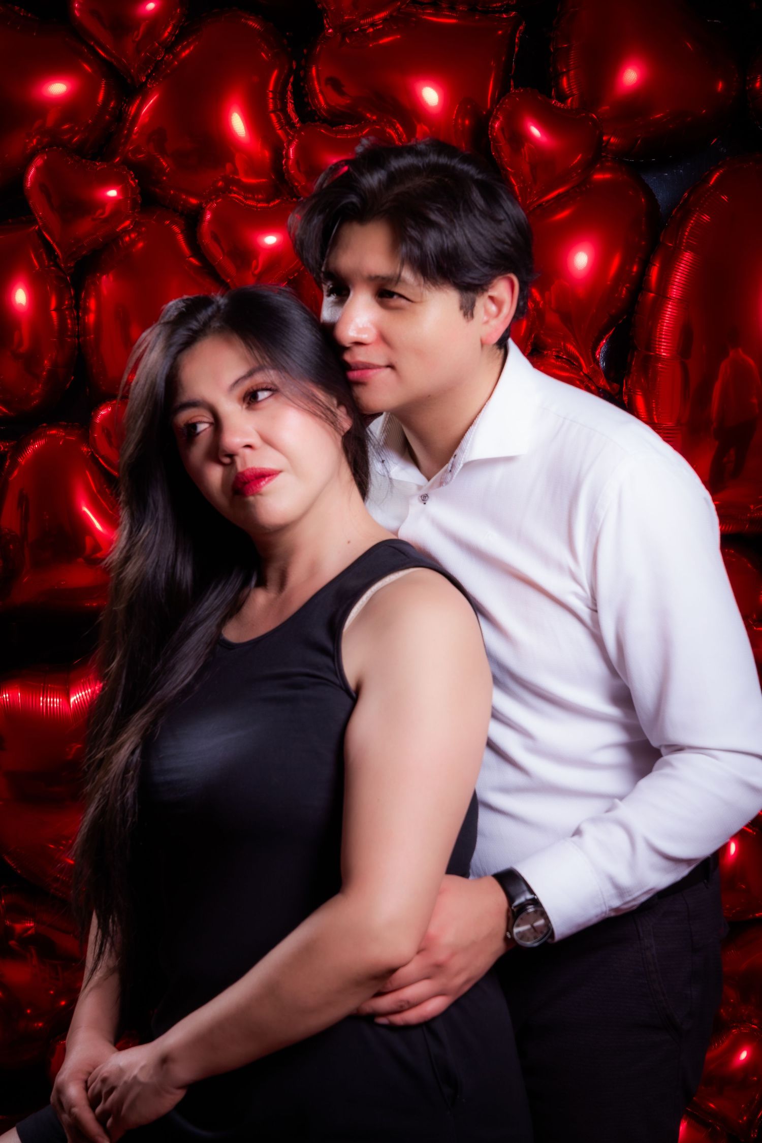 Couple poses intimately against red glowing heart lights in romantic portrait setting.