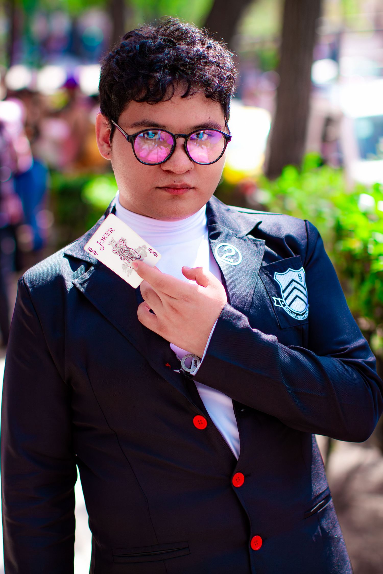 A formal portrait of someone in a black suit with red buttons and round glasses holding a card.