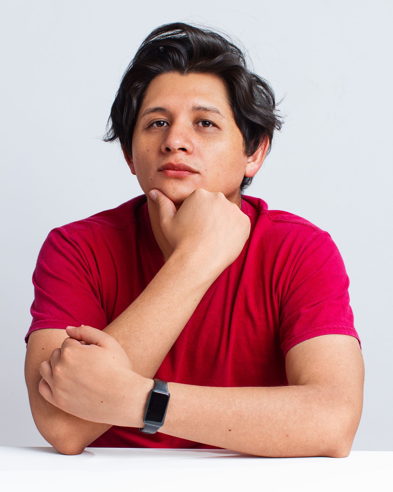 Person in red t-shirt with dark hair poses thoughtfully with hand on chin against white background.
