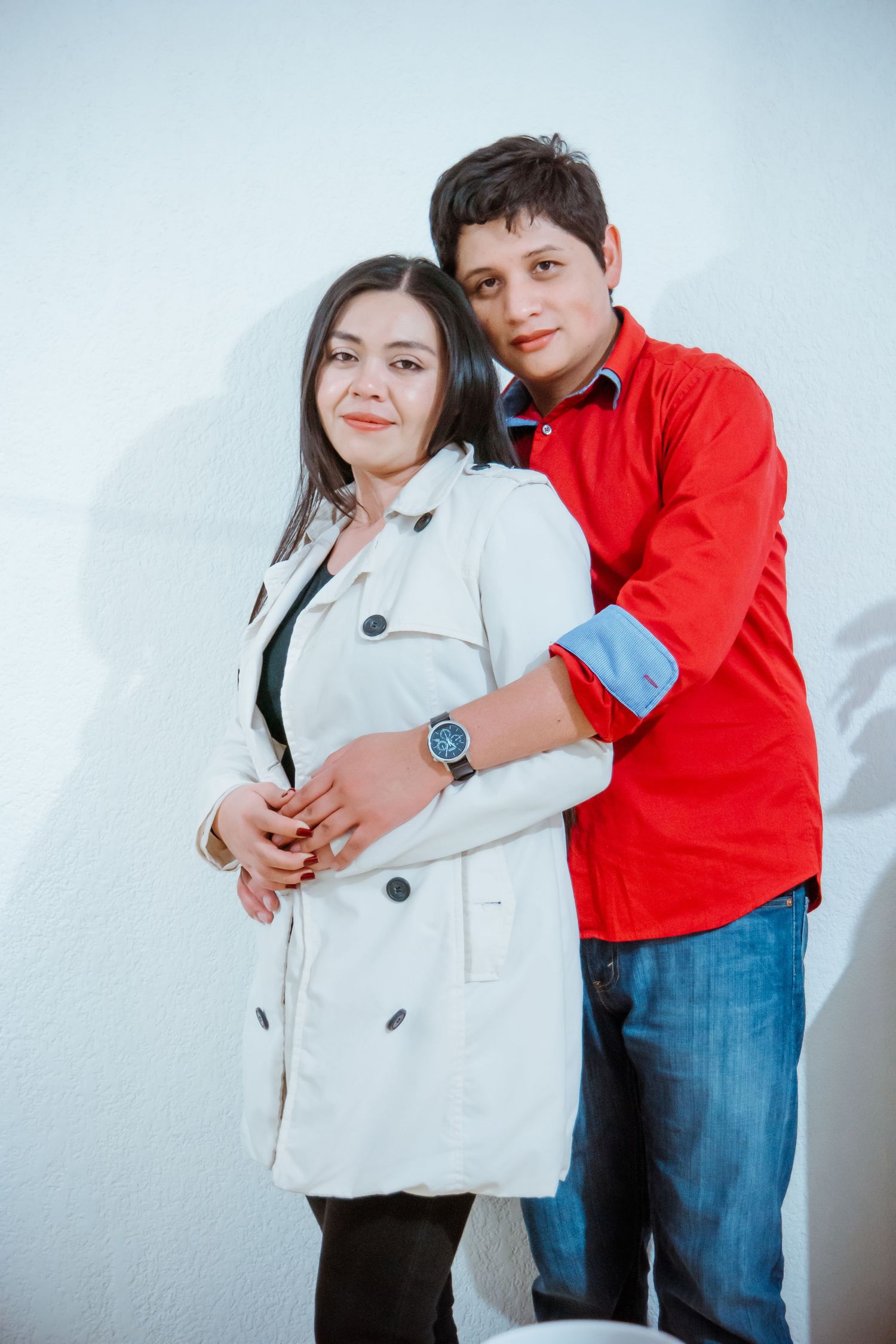 A couple poses together against a white wall, wearing a white coat and red shirt with jeans.