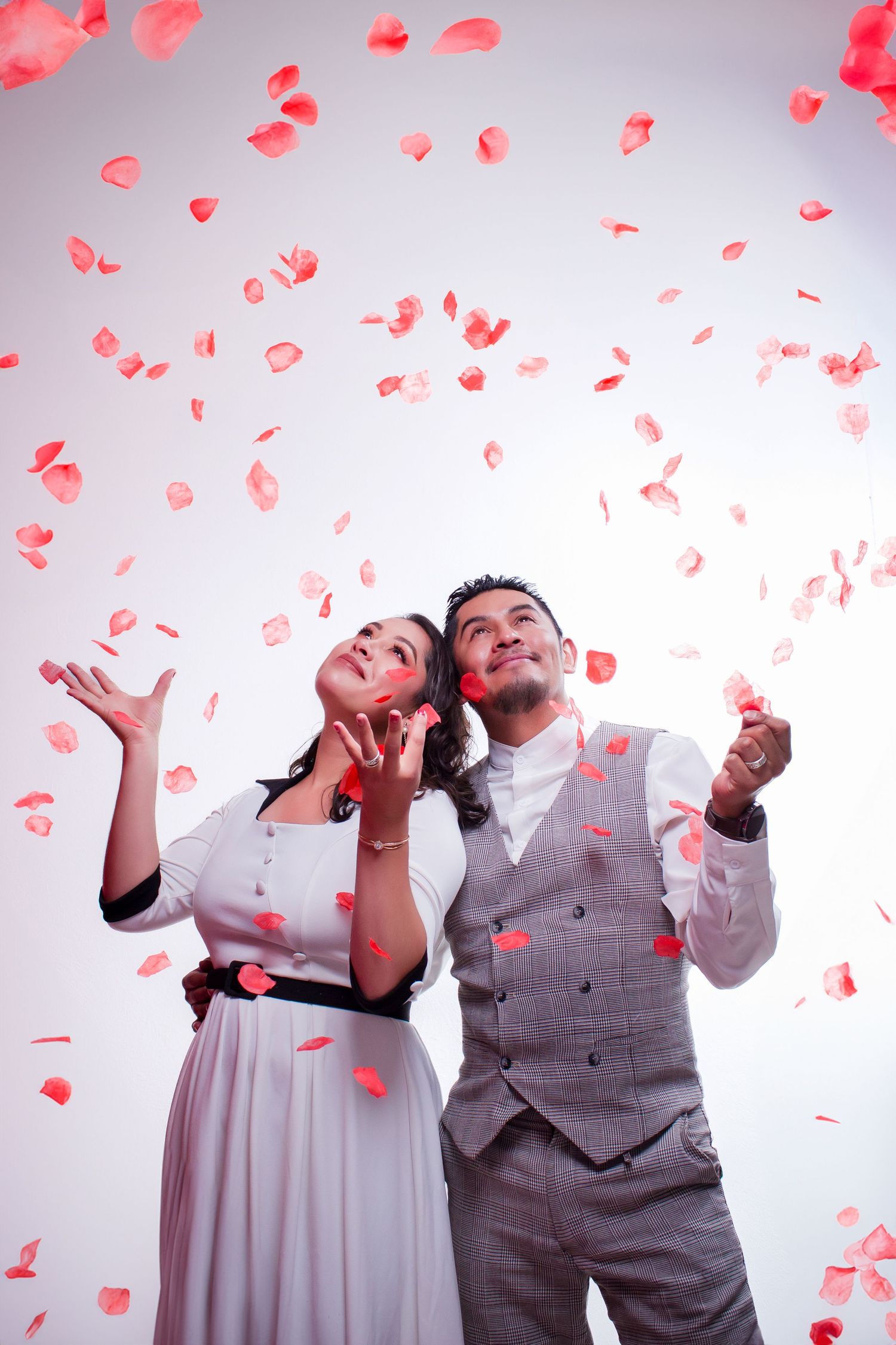 Couple celebrates with falling red rose petals against white background in romantic photoshoot.