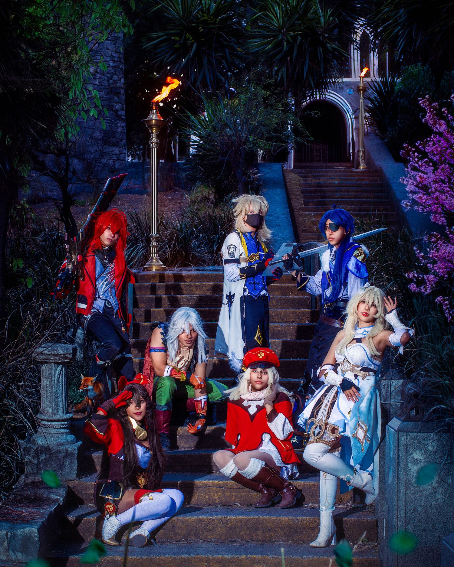 A group of cosplayers in fantasy costumes pose on stone steps at night with glowing lights and gothic architecture.