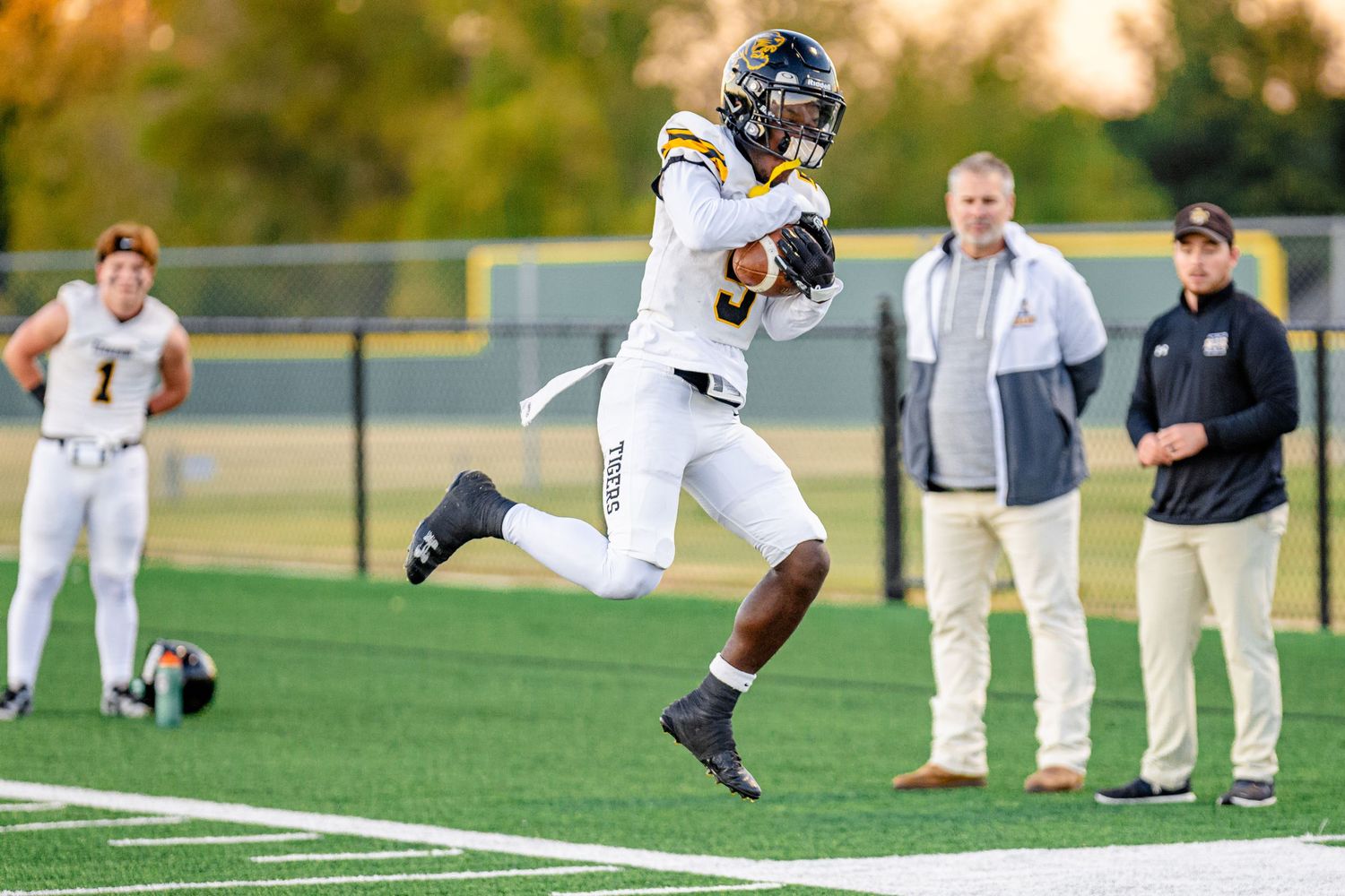 Football player in white and blue uniform leaps to catch the ball during game while coaches watch from sideline.