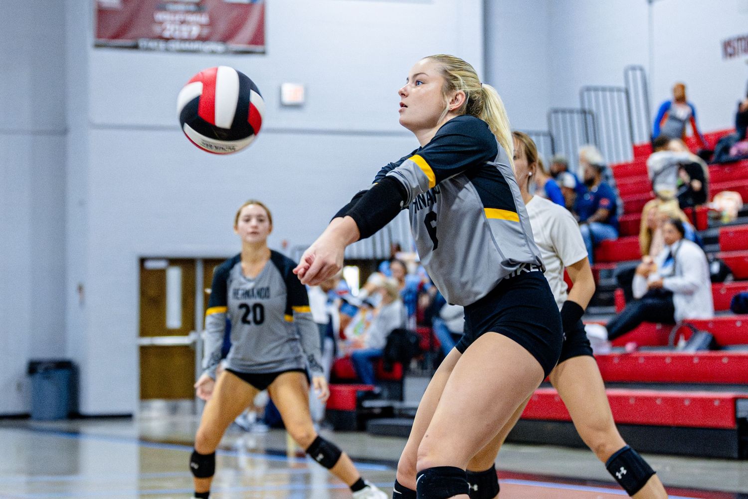 Volleyball player in black uniform lunges to return ball during indoor match with teammate in background.