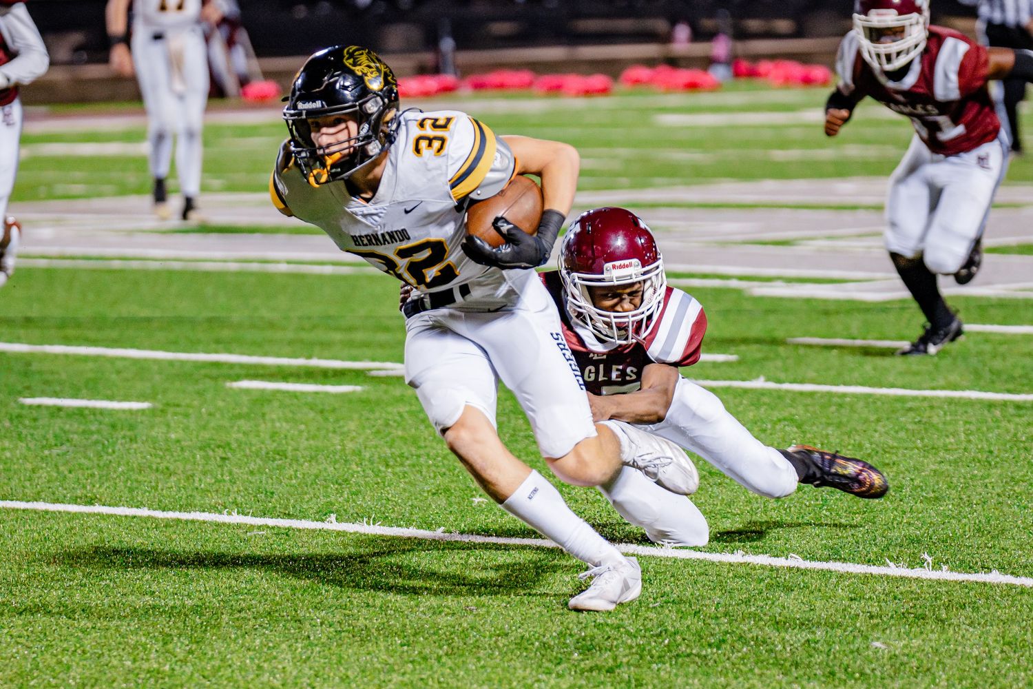 Football player in white and gold uniform being tackled by defender in red and white during night game on turf field.