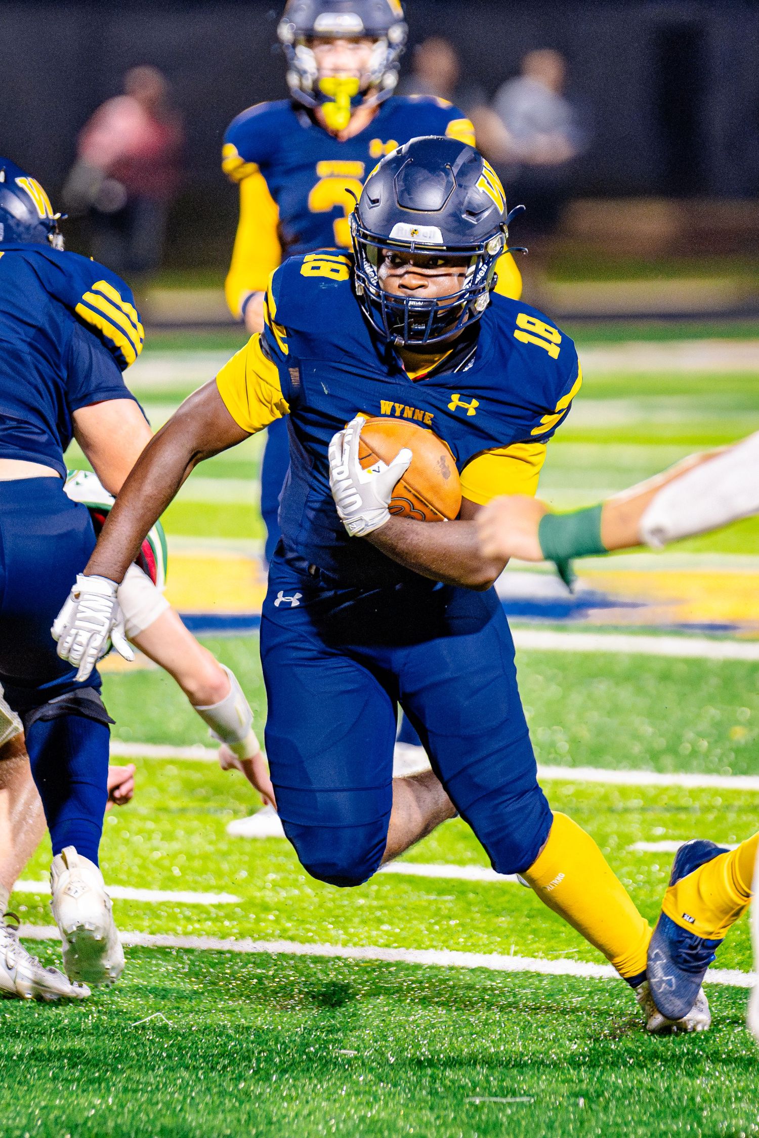 Football player in blue and yellow uniform carries ball while running on field during night game.