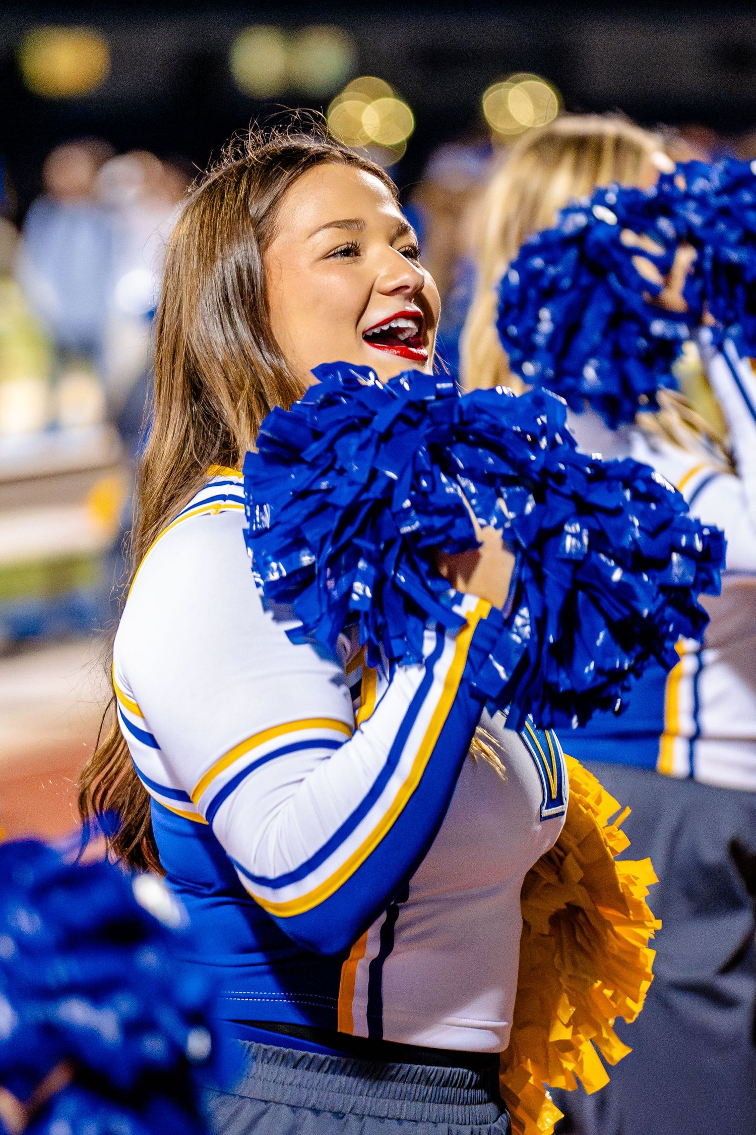 College cheerleaders in blue and gold uniforms perform during a sports event with pom poms and energetic movements.