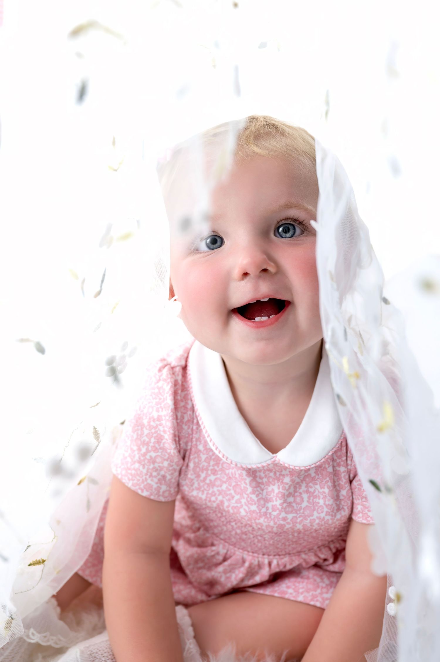 Baby with bright blue eyes and pink dress smiles joyfully under white lace veil.
