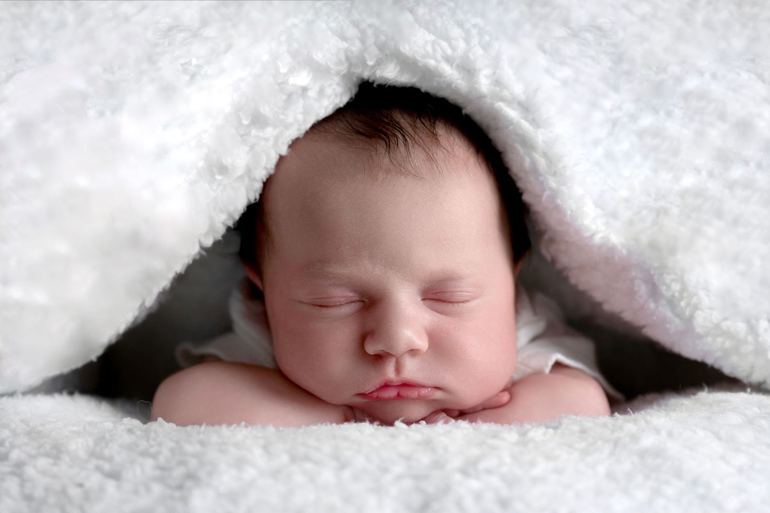 Sleeping newborn nestled under soft white blanket in artistic portrait.