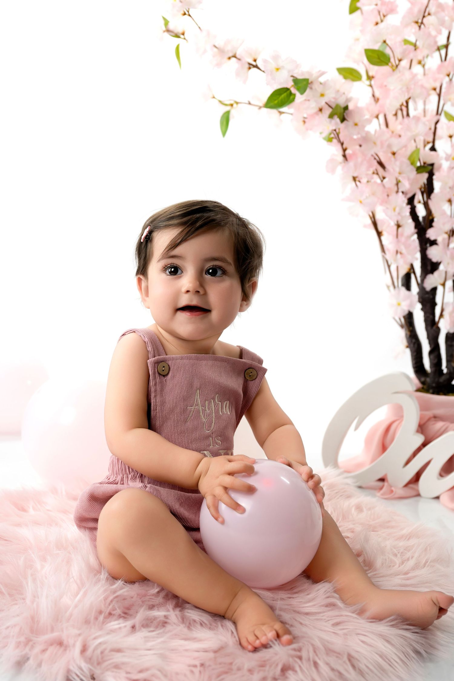 Baby in pink overalls sits on fluffy rug with cherry blossoms and balloon decoration.