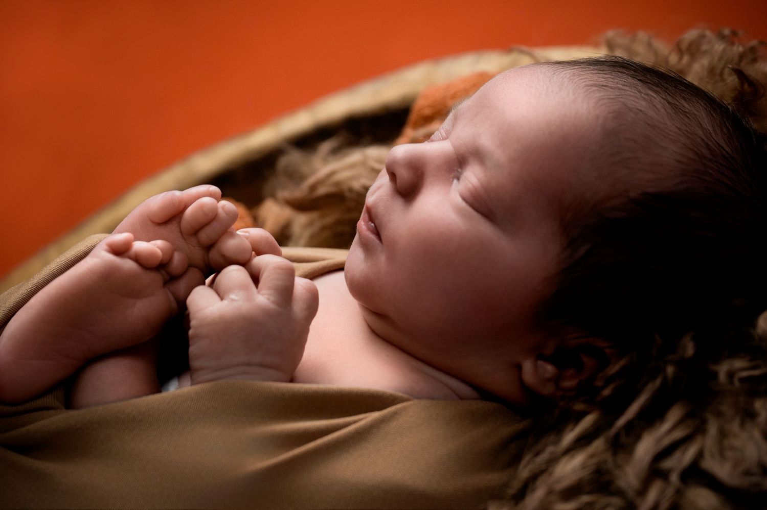 Sleeping newborn on warm brown textured fabric against orange background.
