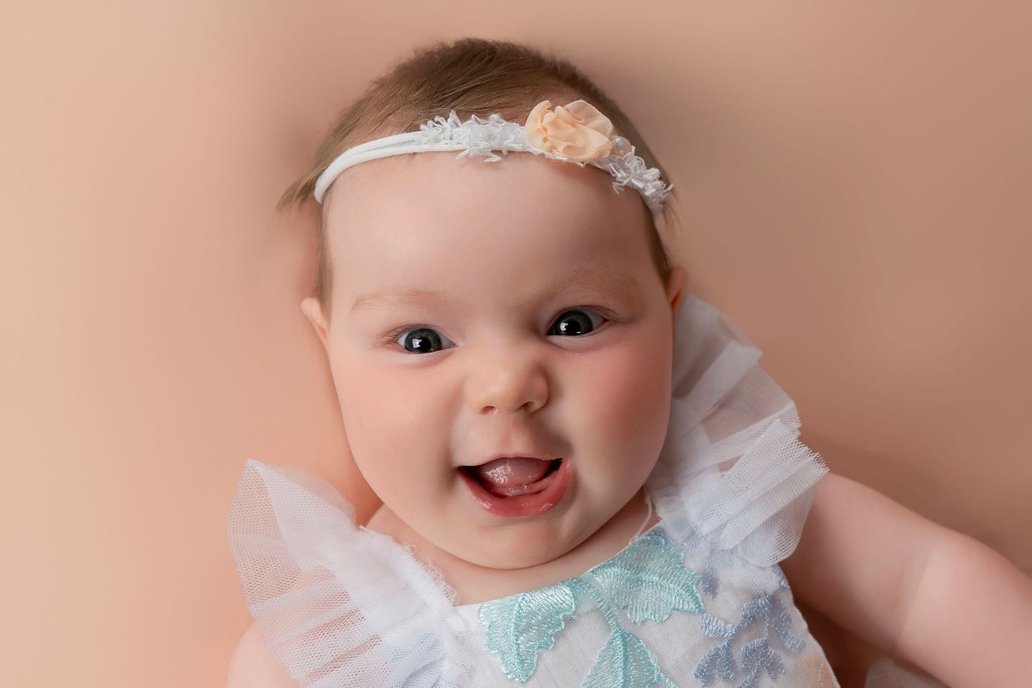 Happy baby wearing white dress and floral headband against pink background.