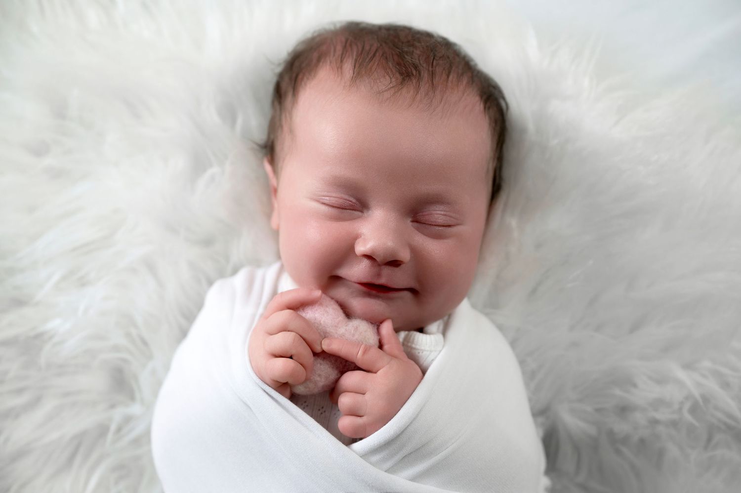 Happy infant wrapped in white swaddle on fluffy white background.