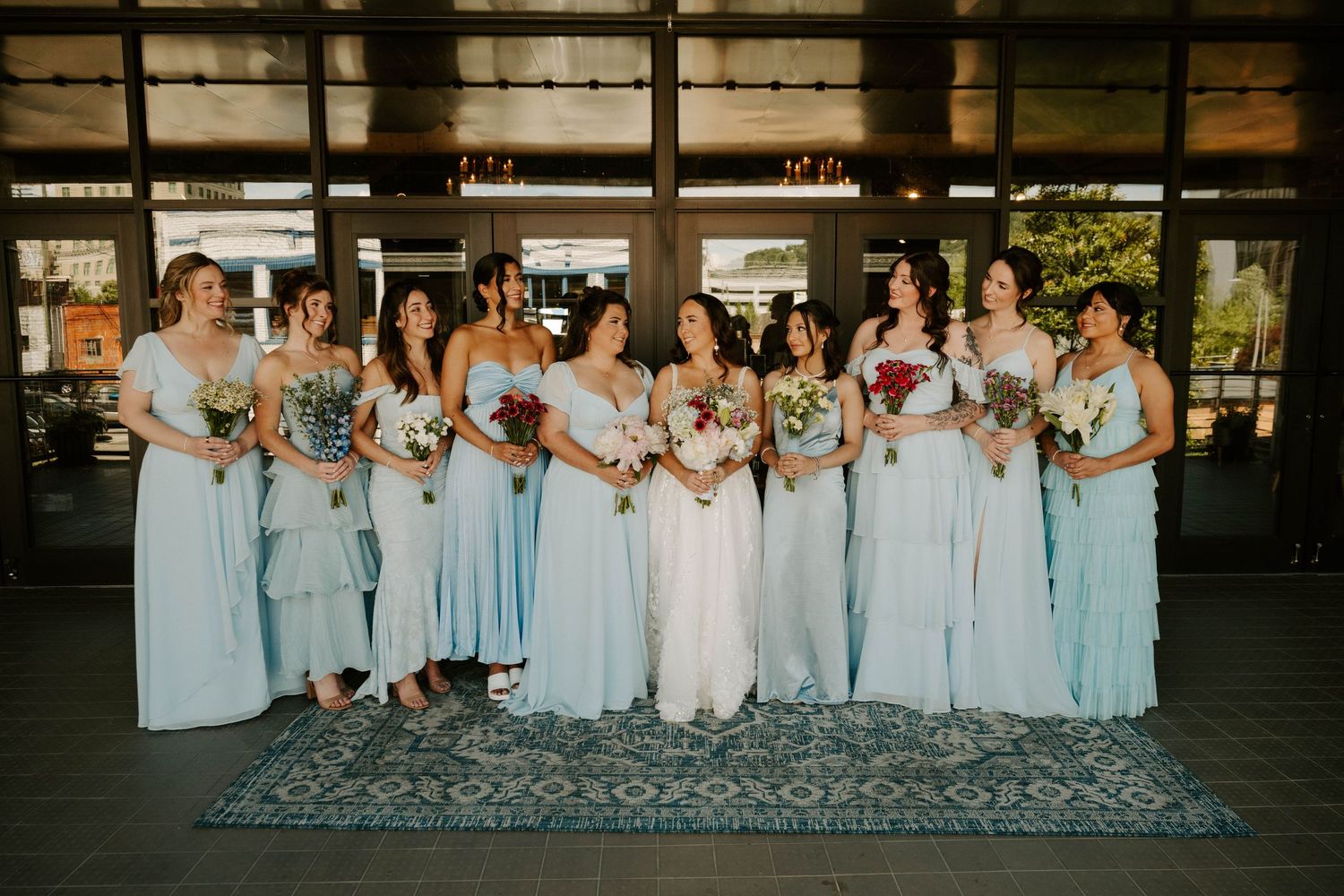 Wedding party wearing pale blue bridesmaid dresses and holding floral bouquets pose together on a patterned rug.