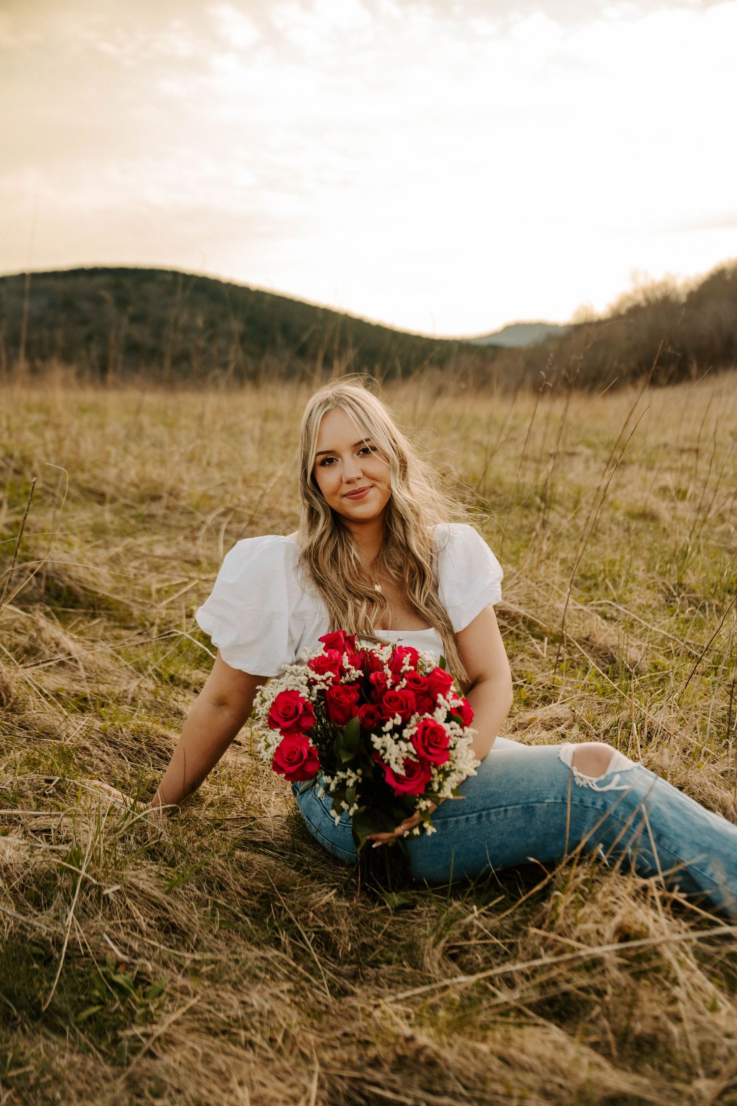 A person in a white top and jeans lies in a grassy field holding a vibrant bouquet of red flowers at sunset.
