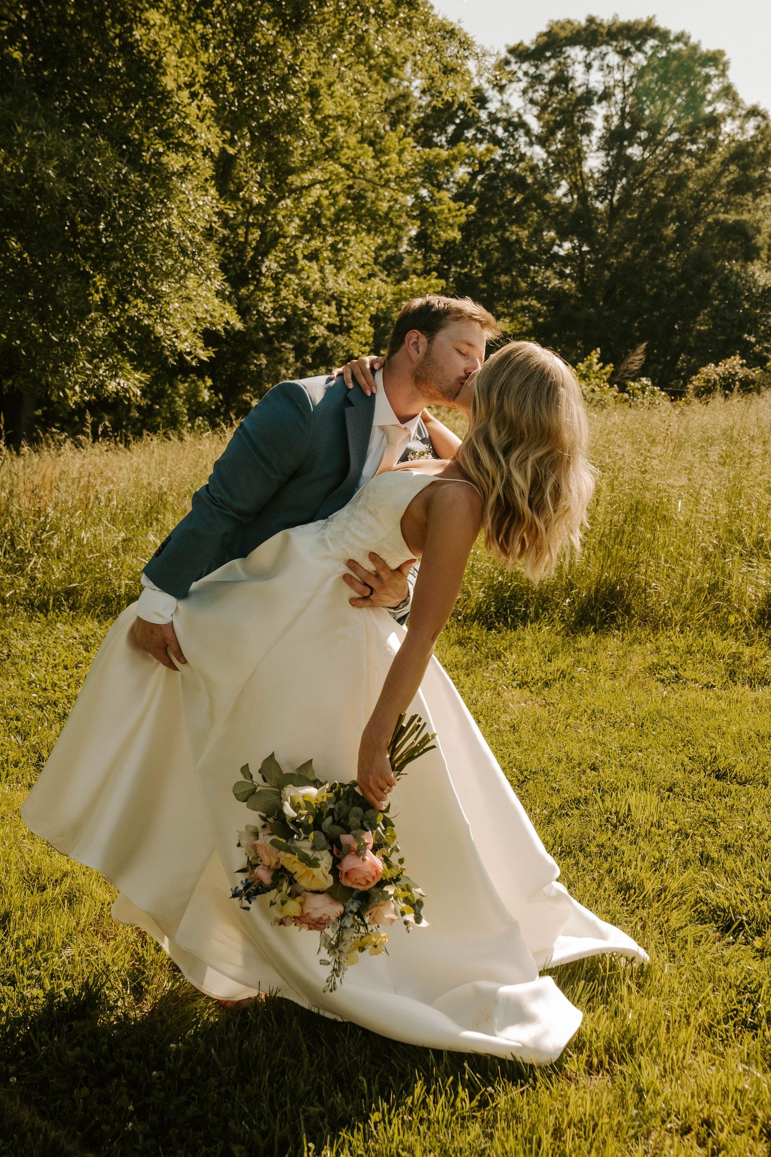Couple in wedding attire share romantic dip pose in sunlit meadow with bouquet at golden hour.