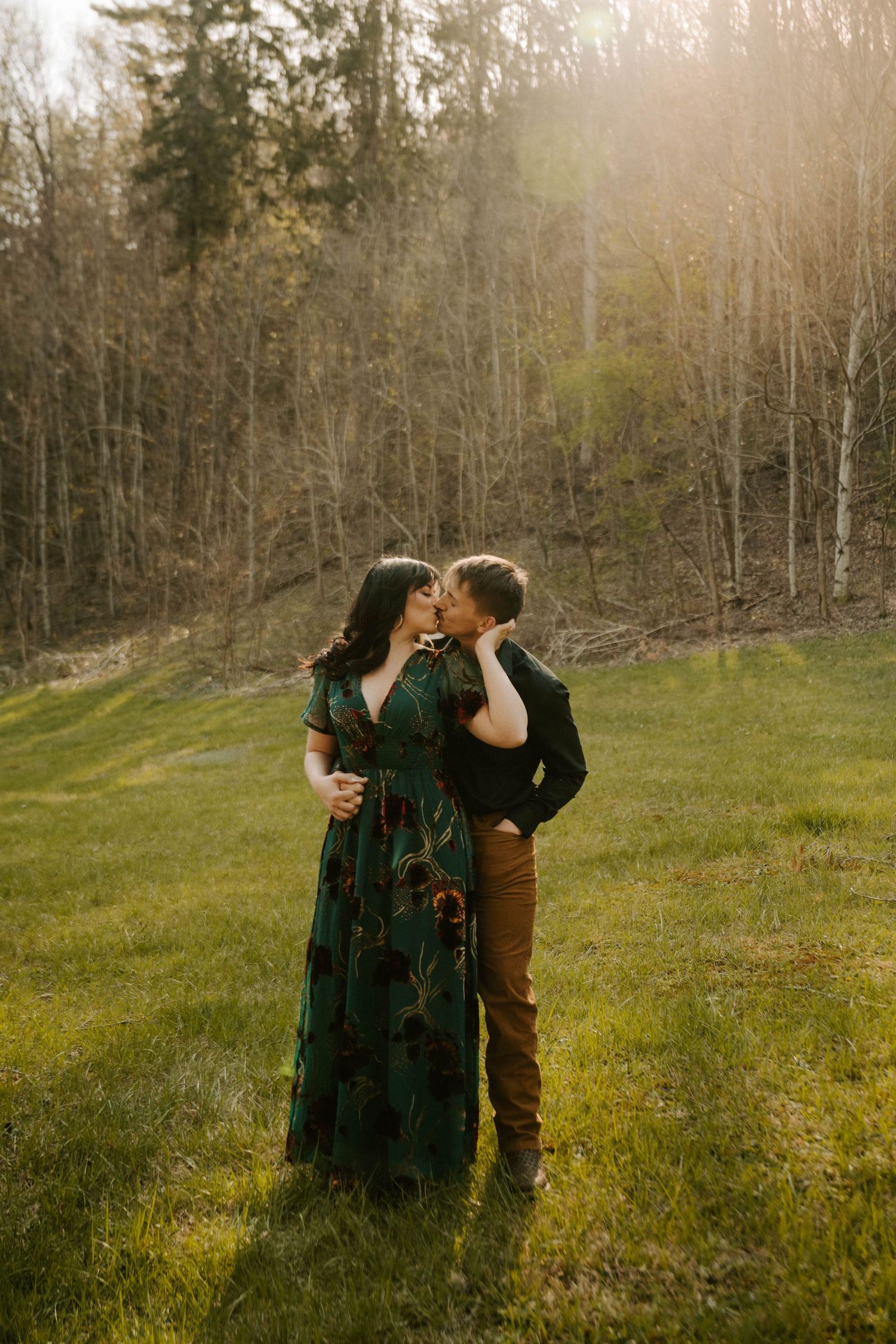 A romantic couple embraces in a sunlit meadow against a backdrop of bare trees during golden hour.