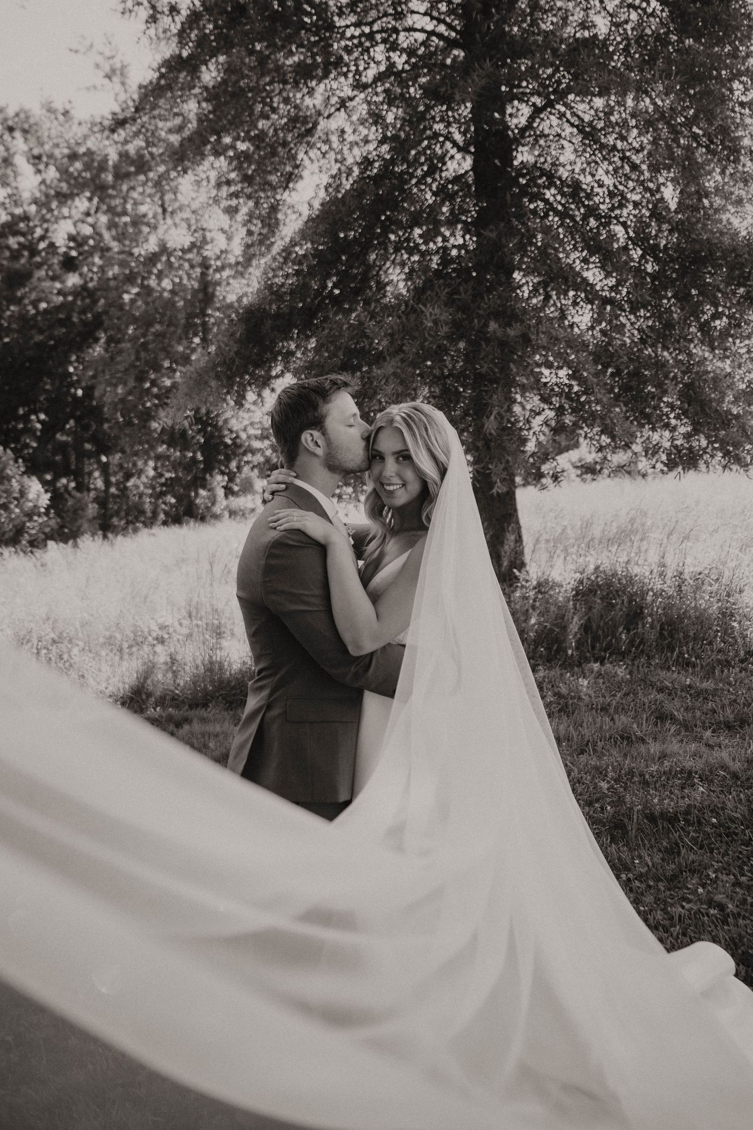 A romantic black and white wedding photo shows a couple kissing under trees while the bride's long veil flows in the breeze.