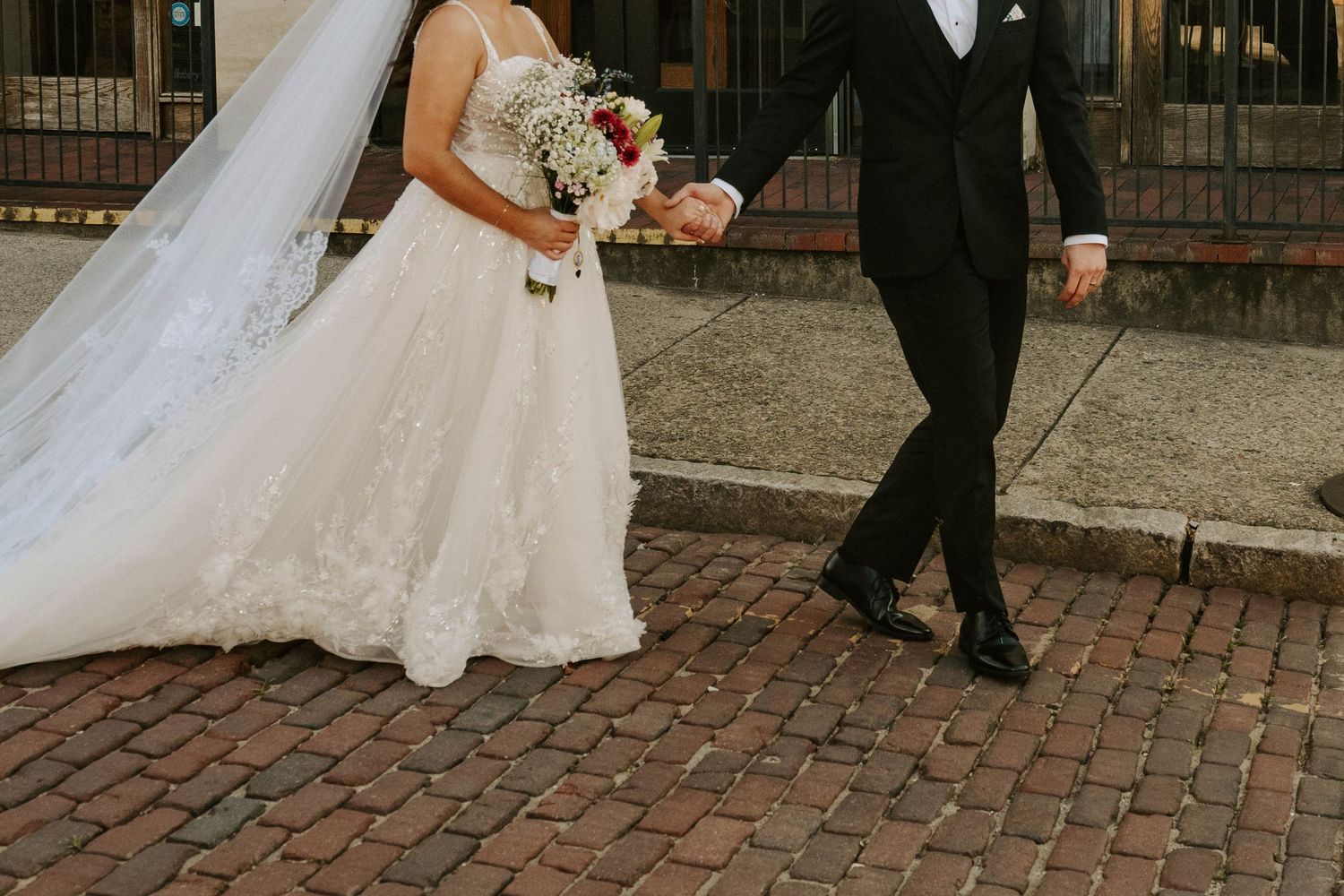 A close-up view of newlyweds walking on cobblestone streets, with bride in white dress holding floral bouquet.